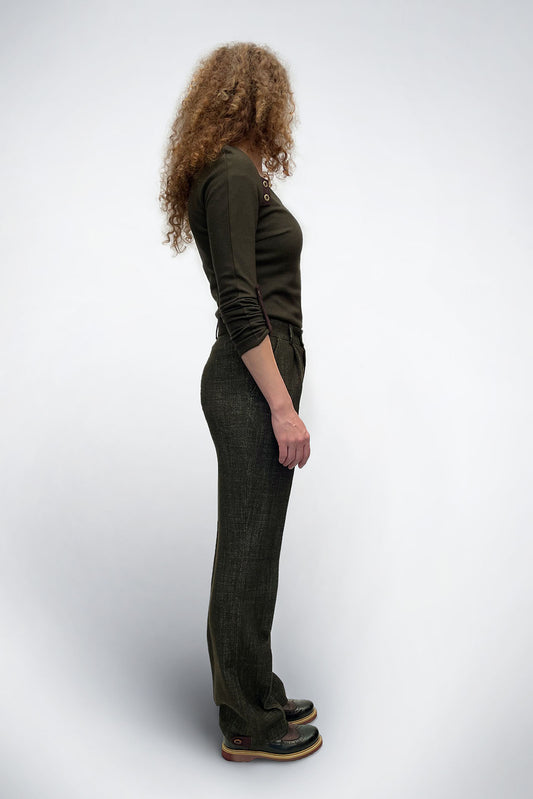 A side profile of a woman with long curly hair wearing the olive tailored trousers. The photo emphasizes the clean, long lines of the straight-leg cut and the high-rise waist, showing how the trousers drape elegantly to the floor.