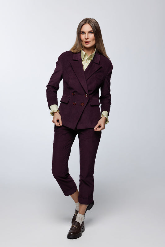 Deep‑purple corduroy double‑breasted suit with cropped tailored trousers and structured blazer, worn by a model with a ruffled blouse and loafers, front studio view.