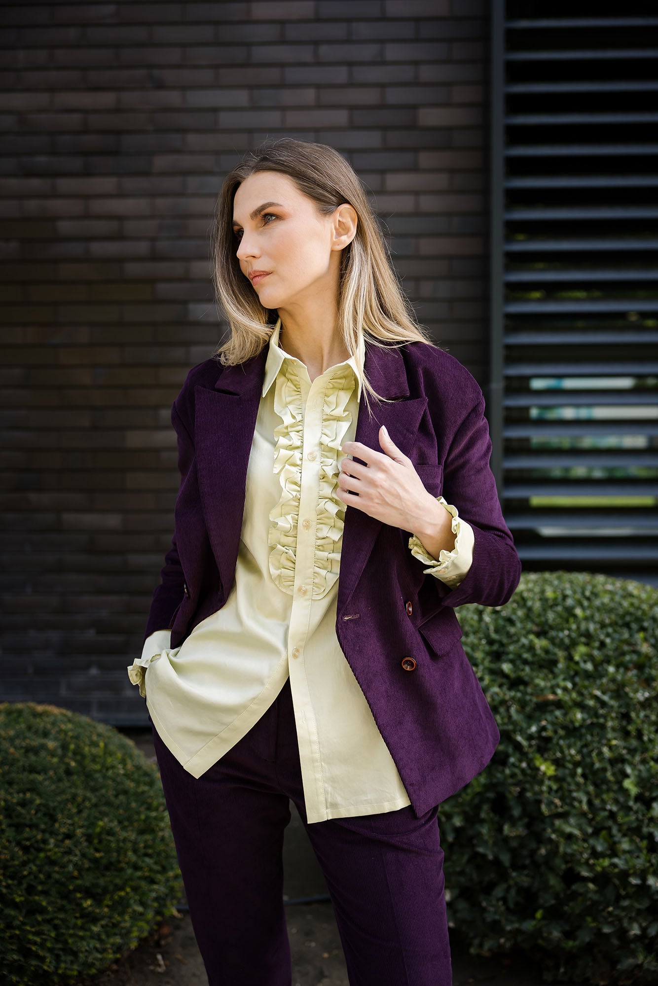 Deep‑purple corduroy double‑breasted suit with structured blazer and matching tailored trousers, worn by a model with a gold‑green sateen cotton blouse with ruffled front and buttoned cuffs, outdoor portrait view.