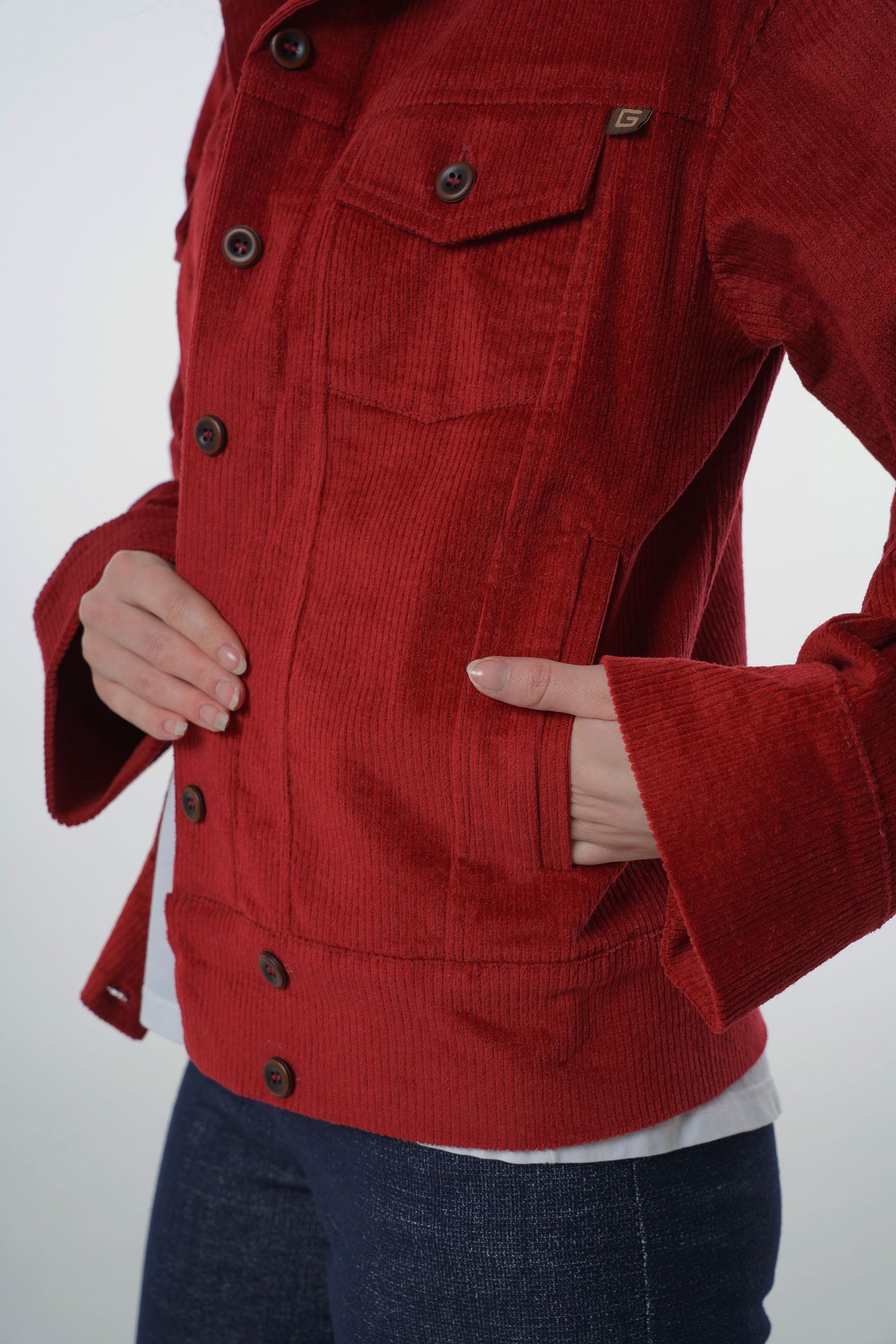 Close-up detail of a dark red corduroy jacket showing the vertical ribbed texture, side welt pocket, and branded logo tag.