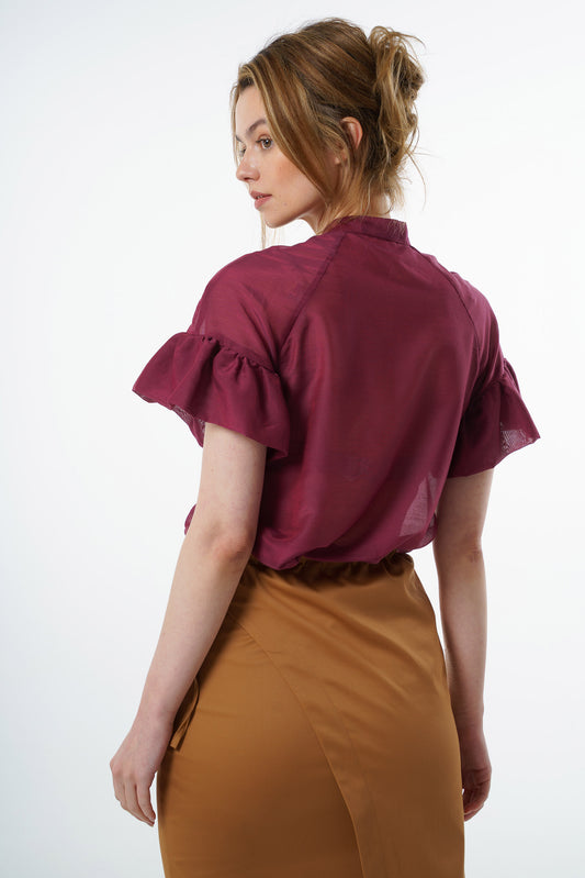 Burgundy viscose–silk raglan‑sleeve blouse with ruffled cuffs and fluid back silhouette, worn by a model with a camel skirt, angled back studio view.
