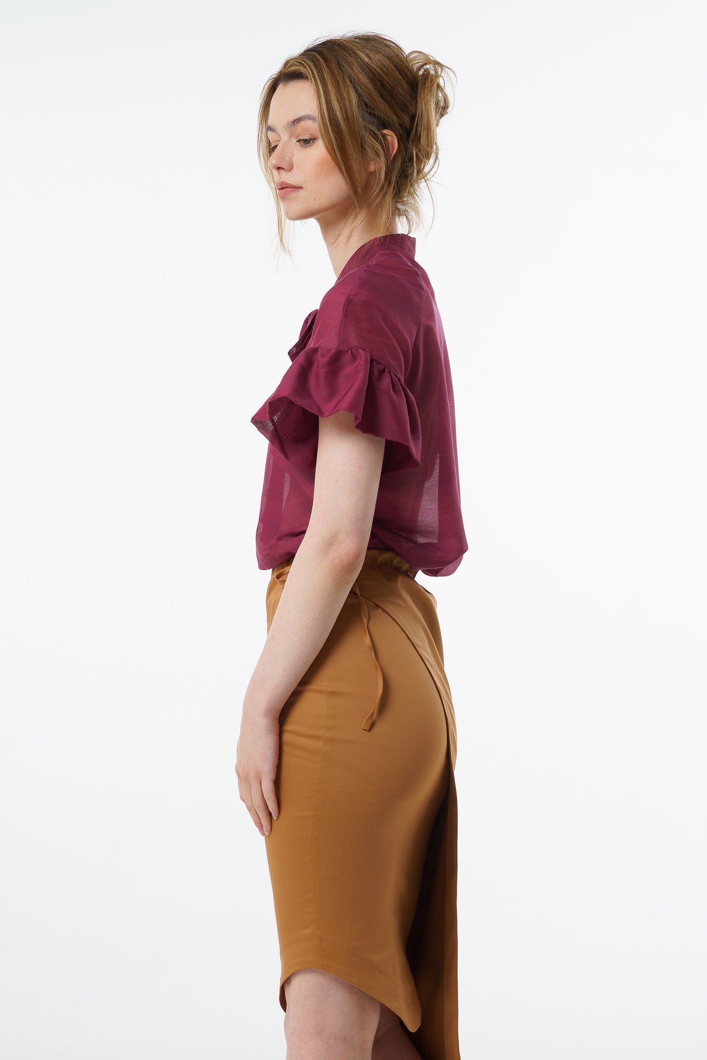 Burgundy viscose–silk raglan‑sleeve blouse with neck tie and ruffled cuffs, worn by a model with a high‑waisted mustard skirt, side studio view.
