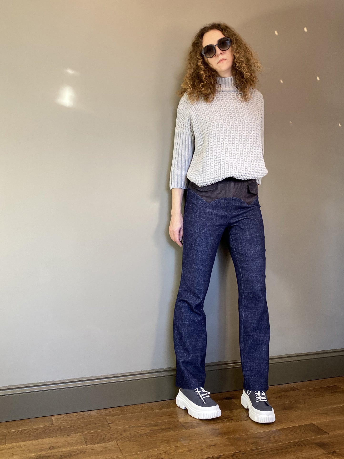 A woman with curly hair poses indoors wearing the high-waisted contrast yoke trousers paired with a chunky grey knit sweater and platform shoes. The textured blue wool fabric stands out against the neutral wall.