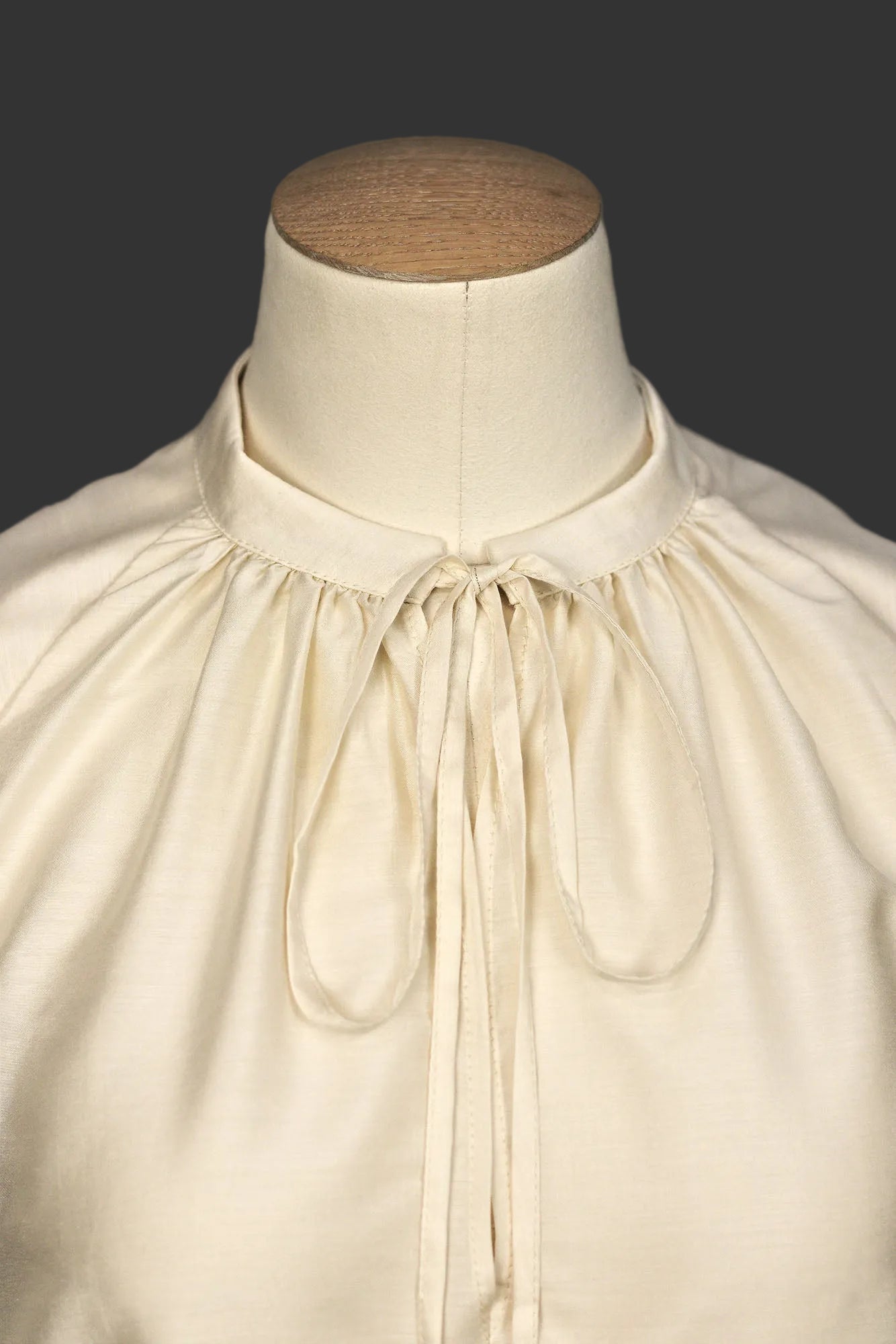 Macro image of ivory sleeveless silk–viscose blouse showing front tie detail, neck gathering and soft sheen, on a mannequin.