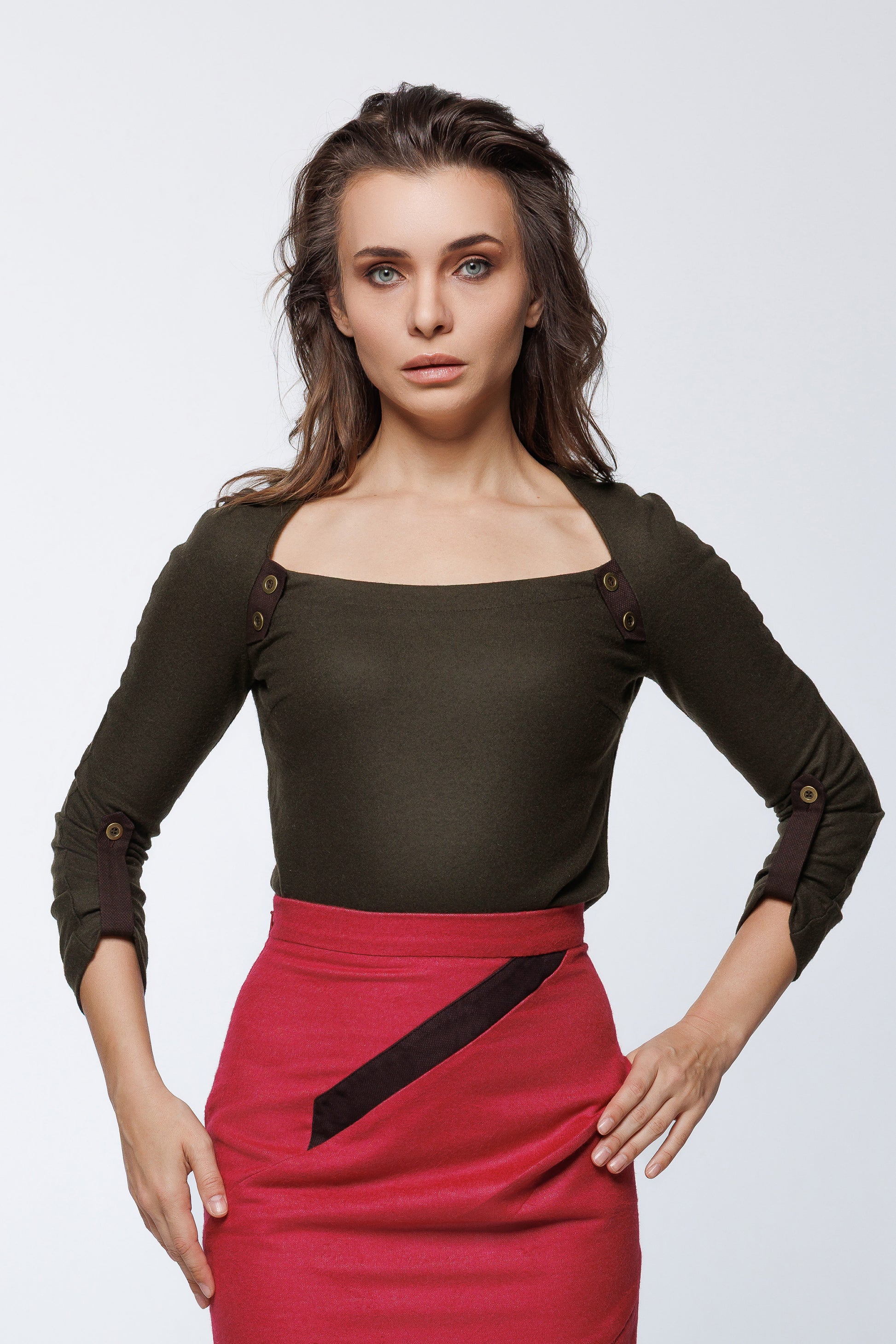 Woman wearing an olive green long-sleeved top with brass button details and a square neckline, paired with a red asymmetrical wool skirt featuring a diagonal accent