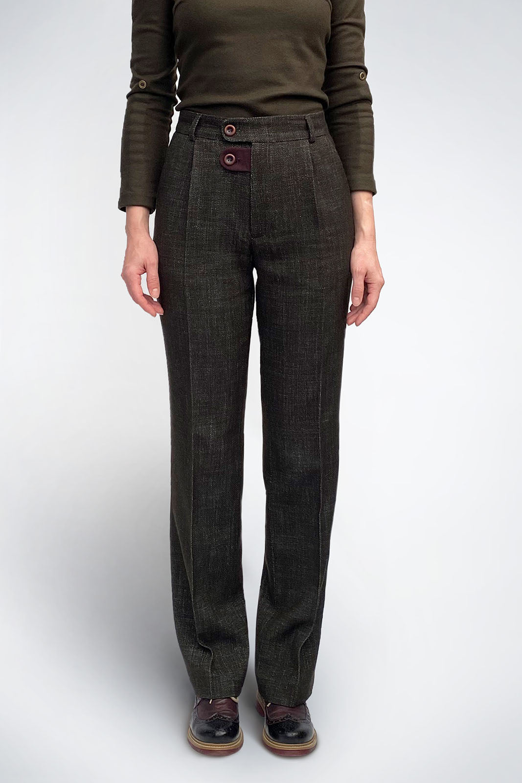 A front view of high-waisted, straight-leg trousers in a deep olive textured wool-cotton blend. The design features a unique double-button closure at the fly with a maroon contrast fabric tab. The trousers are styled with an olive long-sleeve top and burgundy brogues.