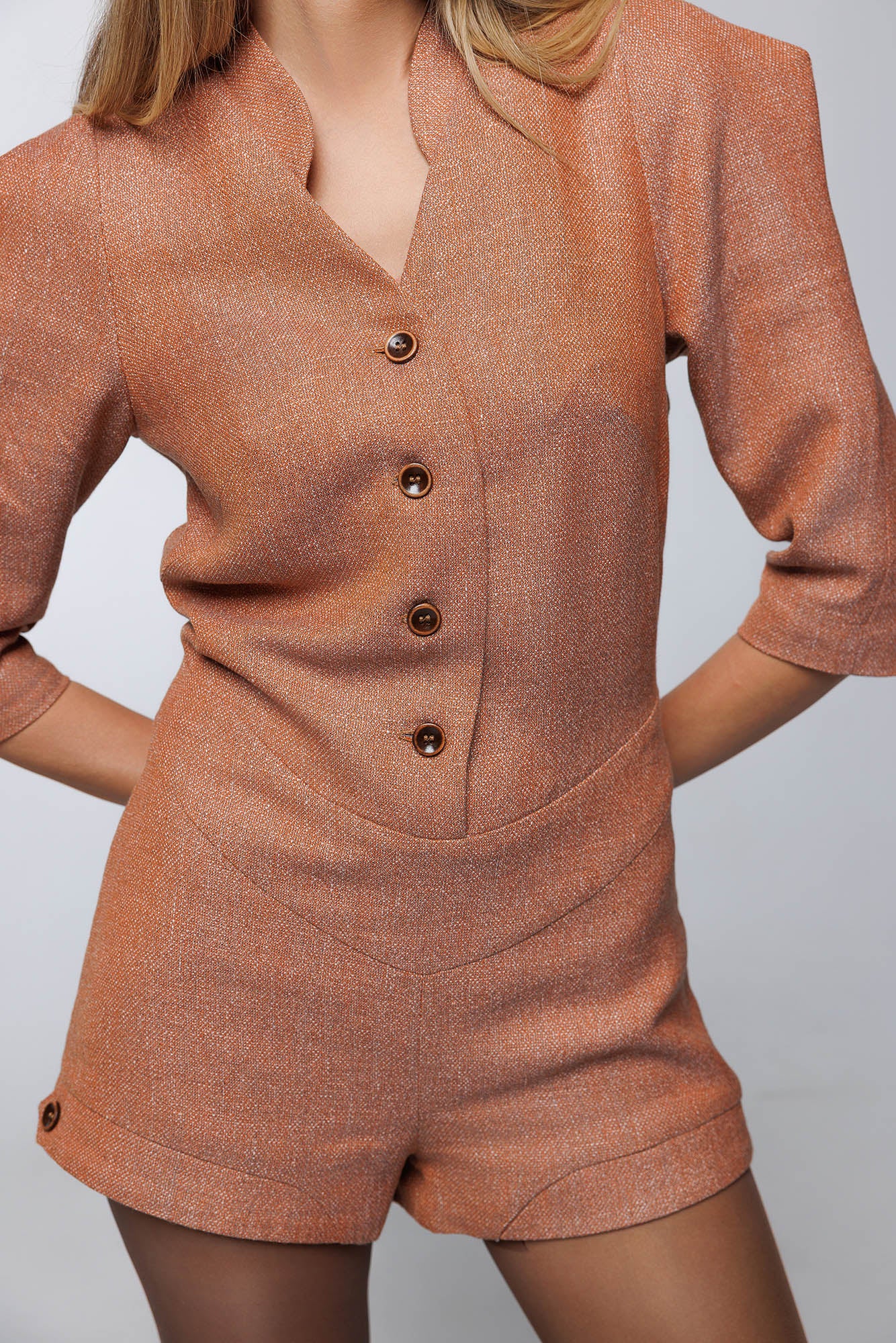 Front close-up of the tailored playsuit showing neckline, button bodice closure, raglan 3/4 sleeves and leg opening buttoned cuffs, emphasizing the fine textured weave of the terracota textured wool-linen fabric.