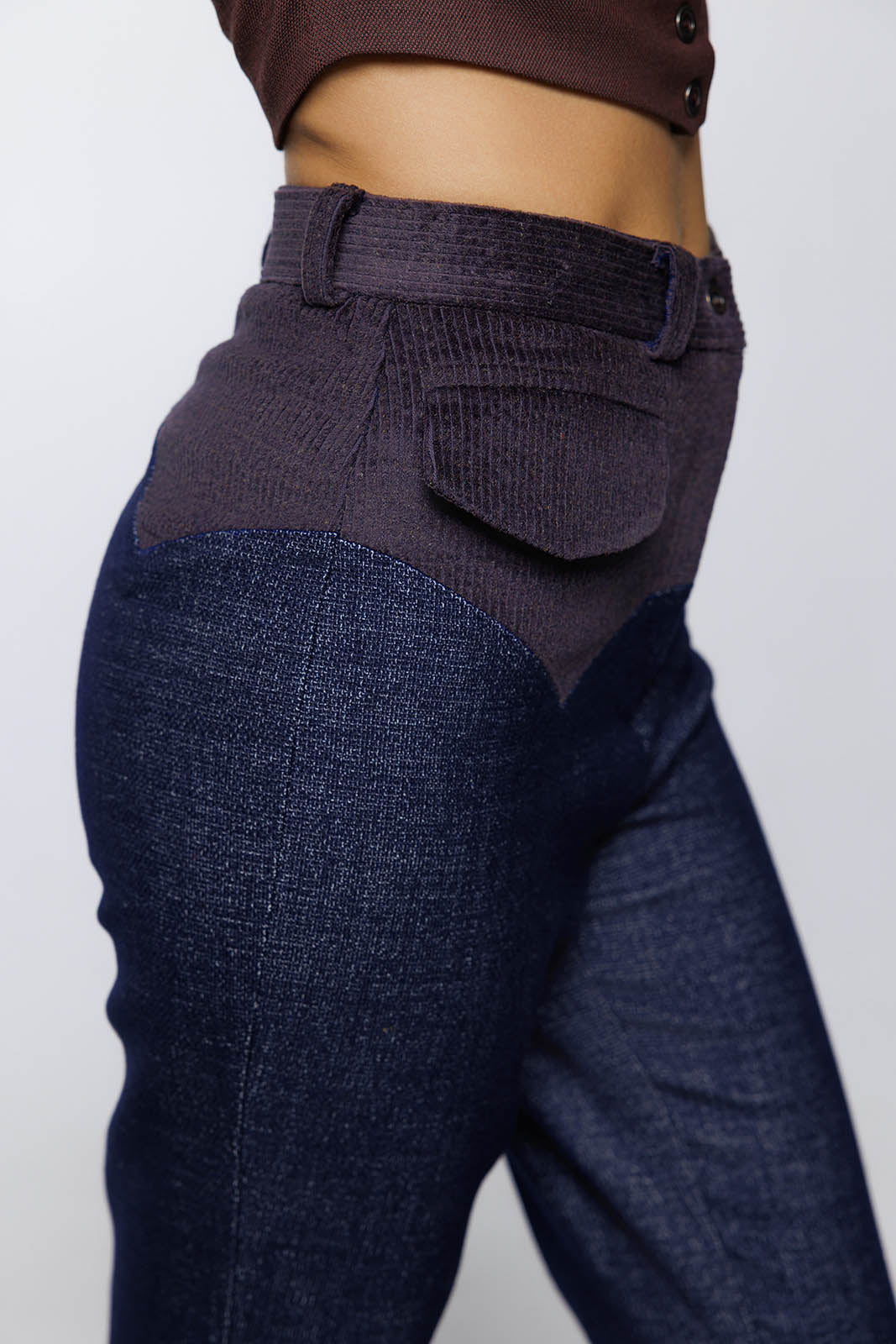 close-up side view of high-waisted trousers featuring a textured blue wool denim body and a dark anthracite corduroy yoke. The yoke has a sharp, pointed "V" shape at the hip, and a small corduroy pocket flap is visible just below the waistband.