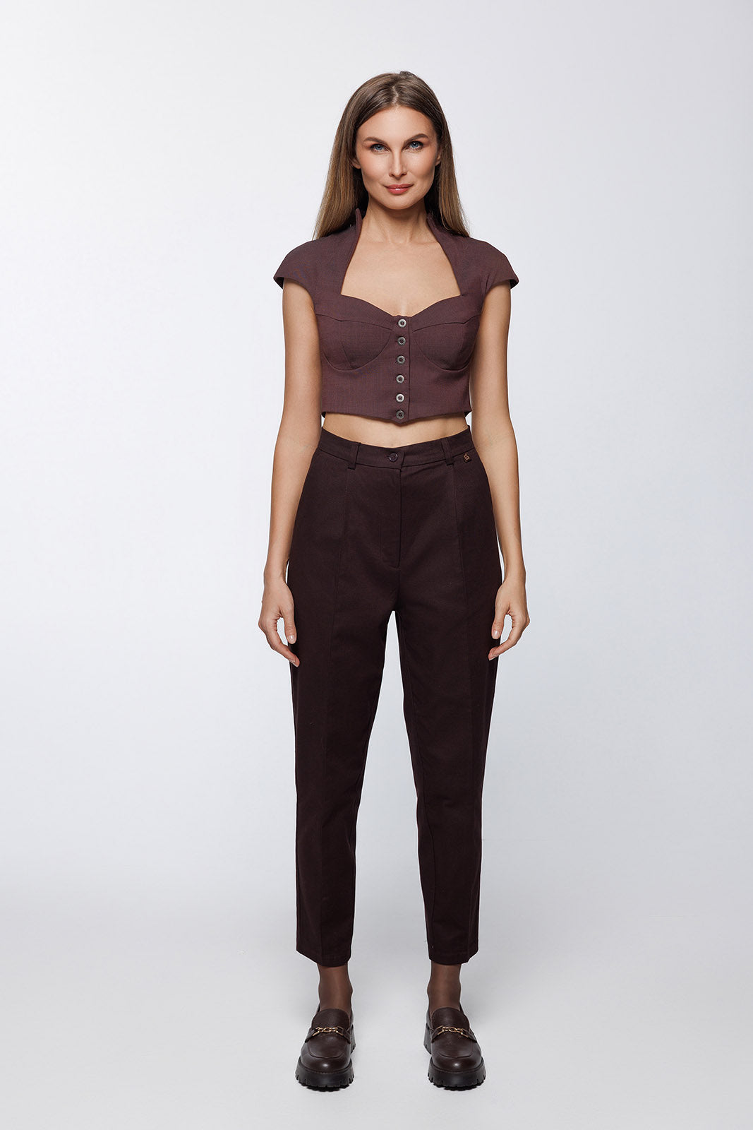 Full-length front view of a woman wearing dark brown tailored trousers with a distinctive vertical seam running down the center of each leg. The trousers feature a high waist, no pleats, and a tapered "carrot" fit ending at the ankle.