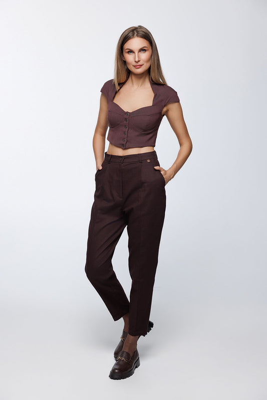Front-angled view of the woman posing with hands in pockets. The image highlights the smooth, pleat-free front of the brown cotton-blend trousers and the sharp central leg seams.