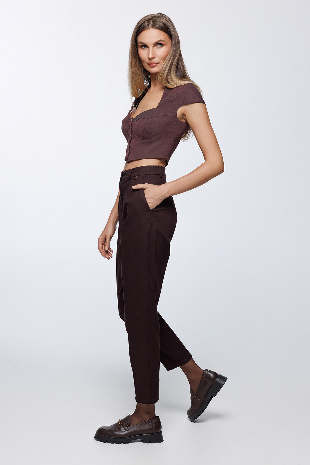 Full-length side profile view showcasing the tapered "carrot" silhouette and the high-waisted fit of the dark brown trousers, paired with chunky brown leather loafers.