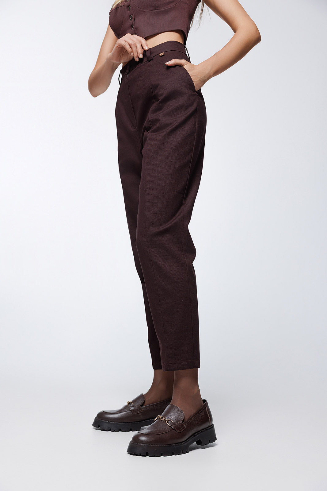 A side-angle shot from the waist down, showing the hand-in-pocket detail and the clean, tapered line of the trouser leg in a dark brown textured cotton blend.