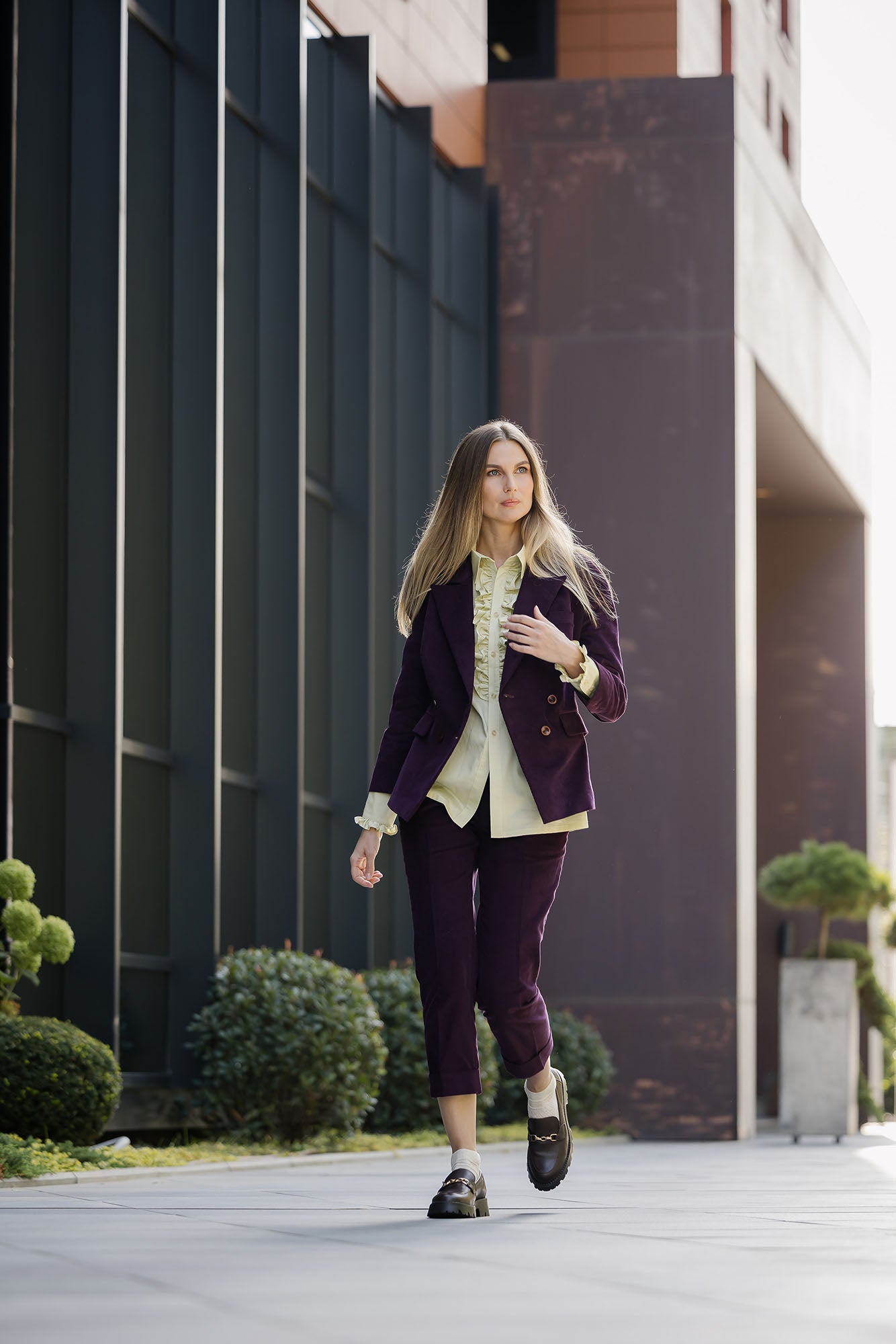 Deep‑purple corduroy double‑breasted suit with structured blazer and cropped tailored trousers, worn by a model with a ruffled blouse and loafers, outdoor walking view.