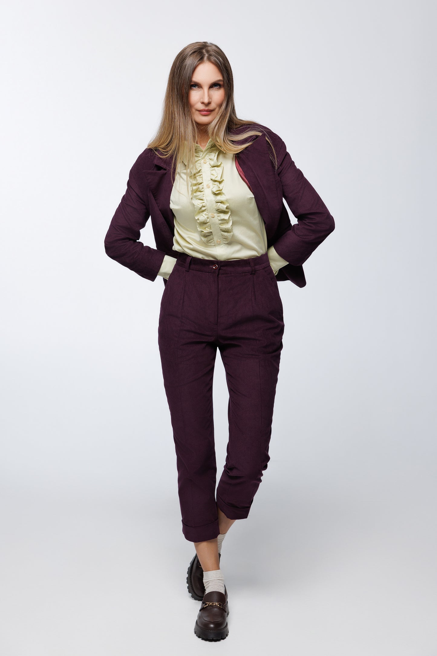 Deep‑purple corduroy double‑breasted suit with tailored trousers and structured blazer, worn by a model with a ruffled blouse and loafers, full‑length studio view.