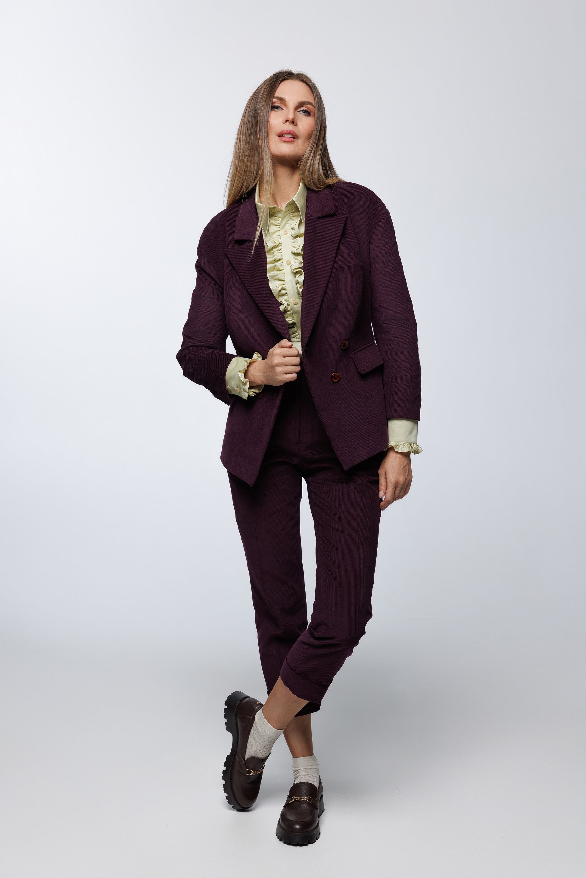 Deep‑purple corduroy double‑breasted suit with matching trousers, worn by a model with a ruffled blouse and loafers, front studio view.