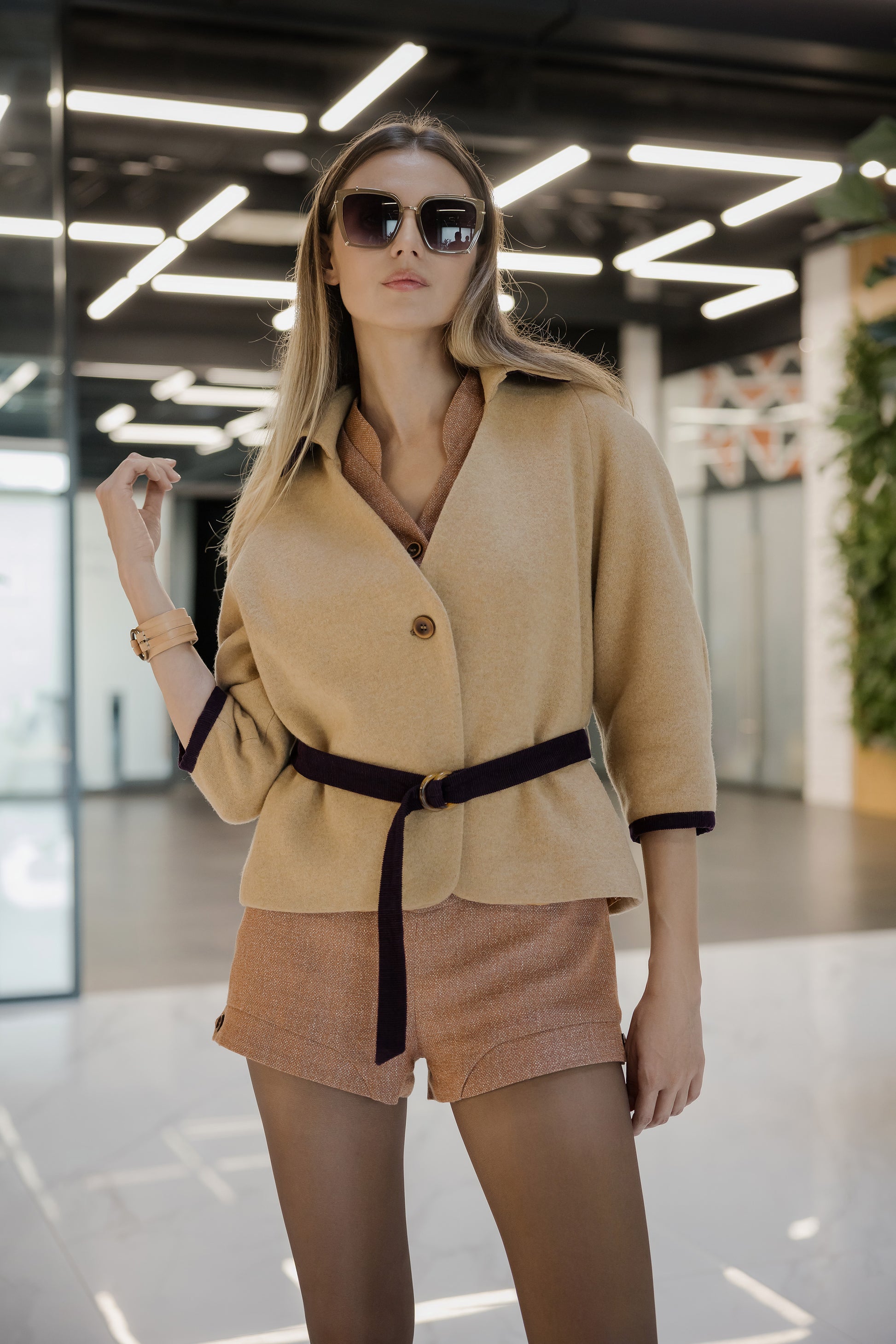 Lifestyle shot of a woman in a honey-beige belted jacket and sunglasses, styled for a modern luxury city look.