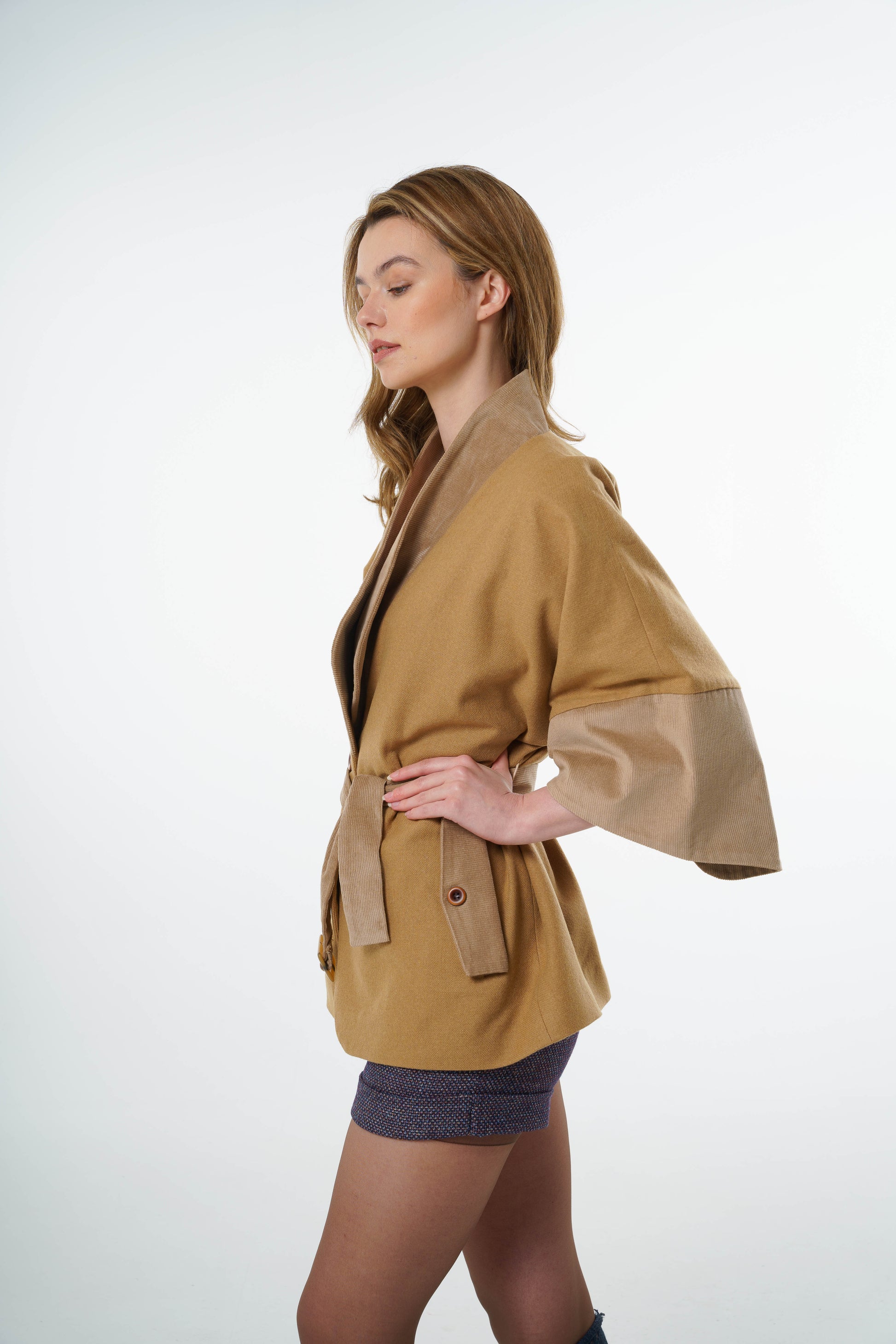 Close-up of a belted jacket worn by a model showing the textured wool-cotton fabric and contrast corduroy cuff detail on a wide sleeve.