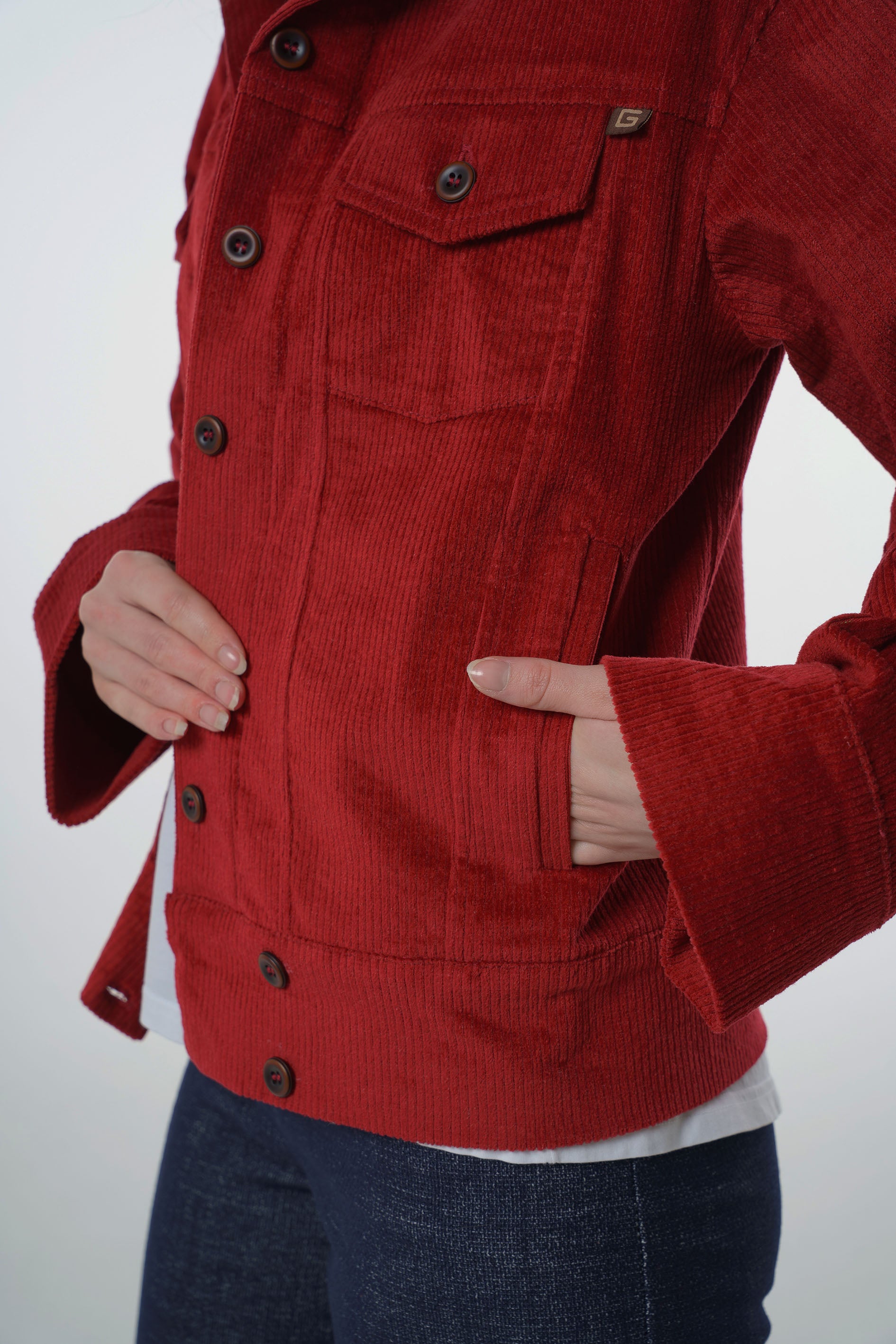 Close-up detail of a dark red corduroy jacket showing the vertical ribbed texture, side welt pocket, and branded logo tag.