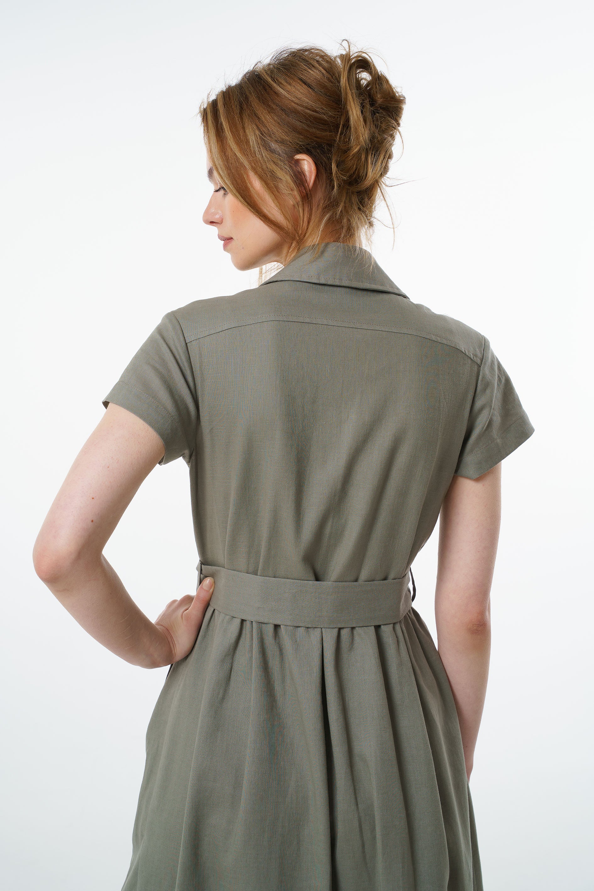 Back view of olive green linen belted dress showing pleated waist and flared skirt, worn by a model.