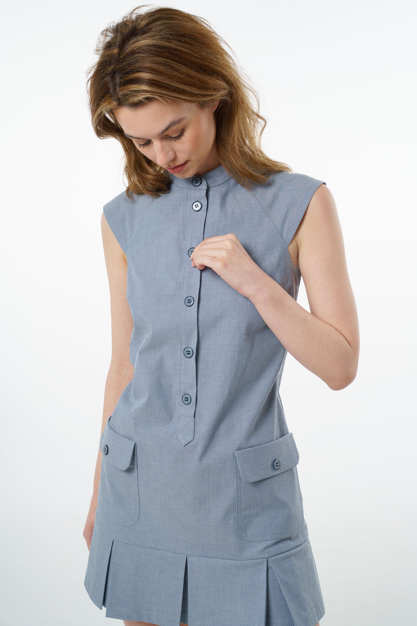 Slate blue sleeveless cotton mini dress with button‑up front, high collar, and flap pockets, worn by a model, close‑up studio view.