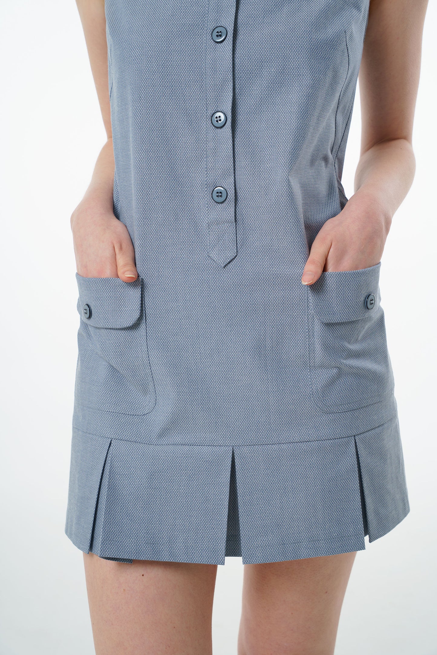 Close‑up of slate blue sleeveless cotton mini dress showing button‑up front, flap pockets, and pleated hem detail.