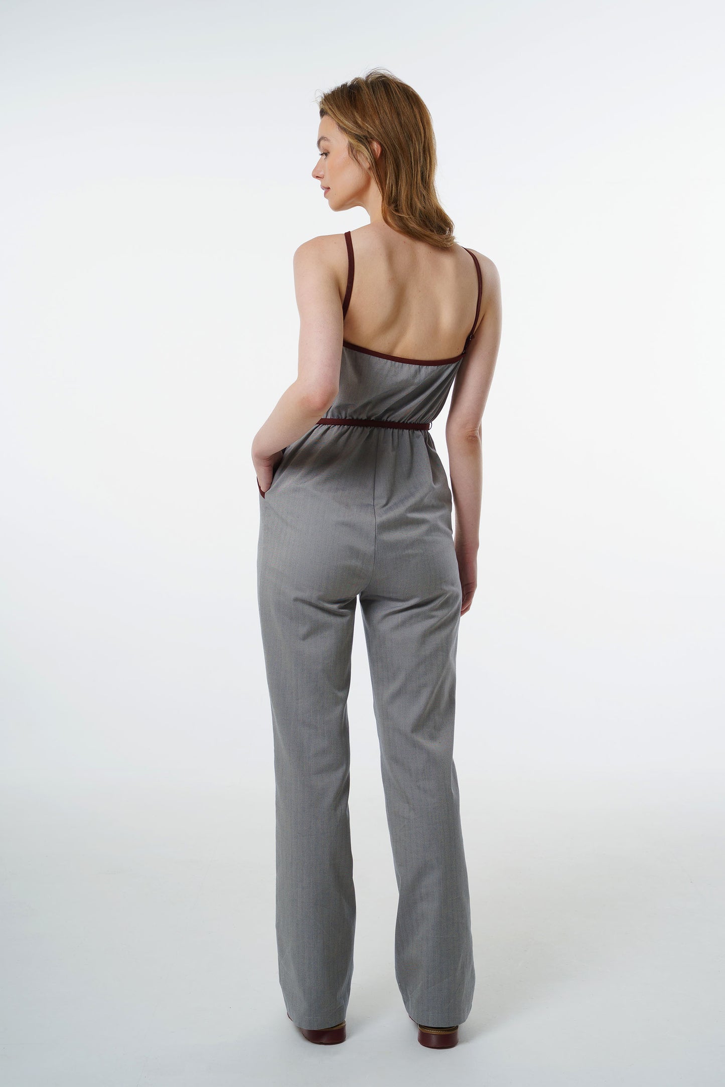 Grey straight‑leg jumpsuit with wine‑red trim and halter straps, worn by a model, back studio view.