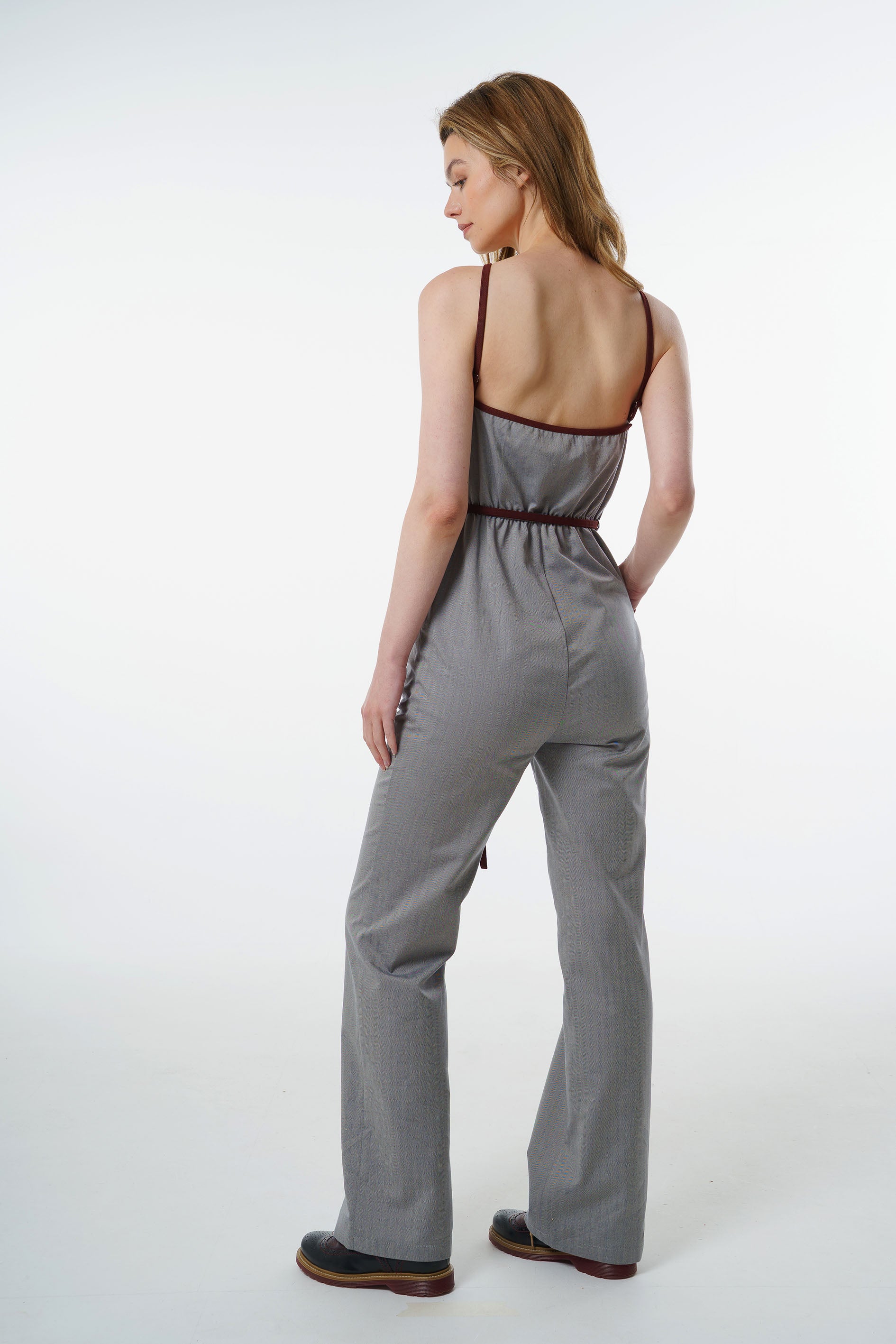 Grey straight‑leg jumpsuit with wine‑red trim and fitted waist, worn by a model, back studio view.