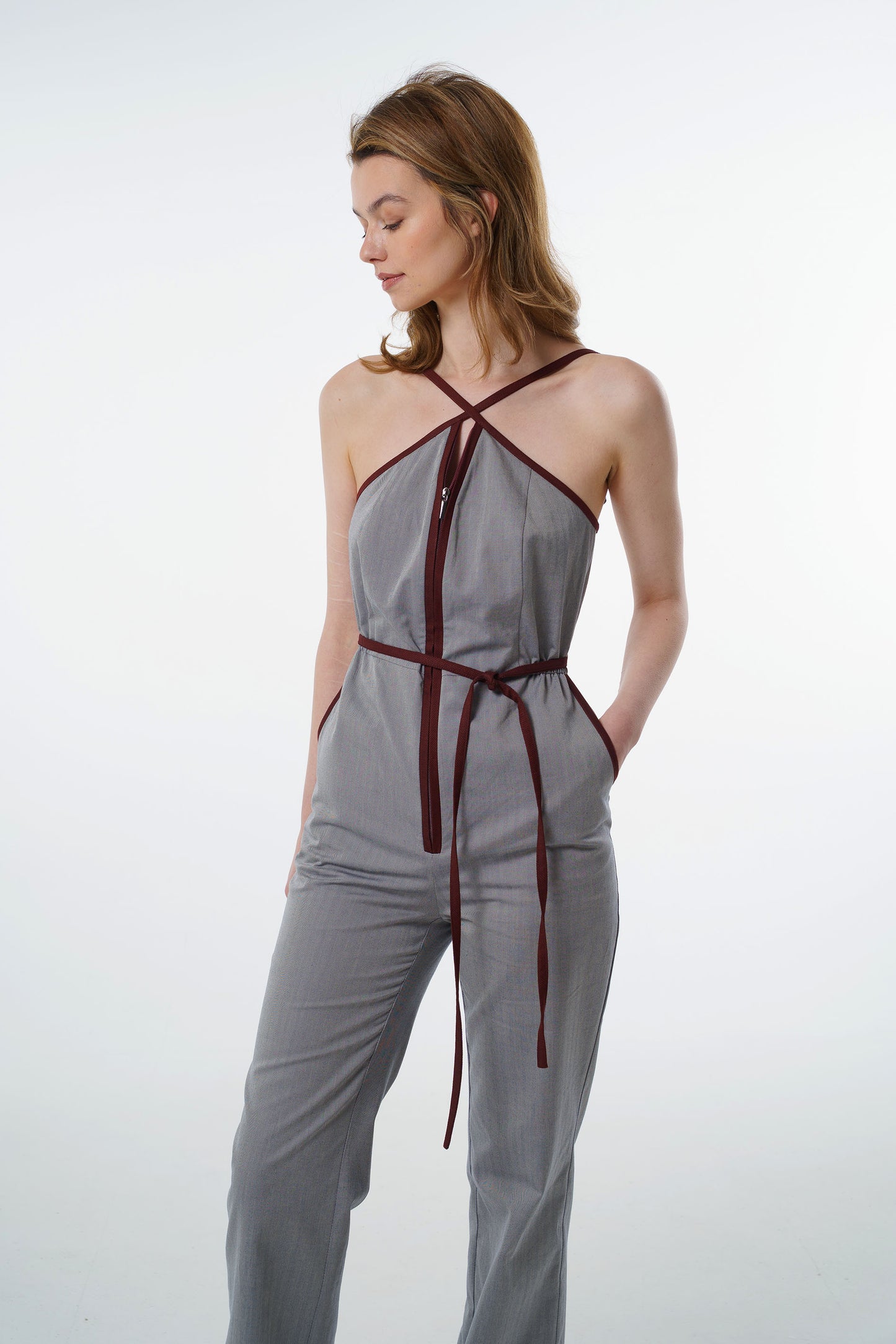 Grey jumpsuit with wine‑red trim, zip‑front closure, and side pockets, worn by a model, front studio view.