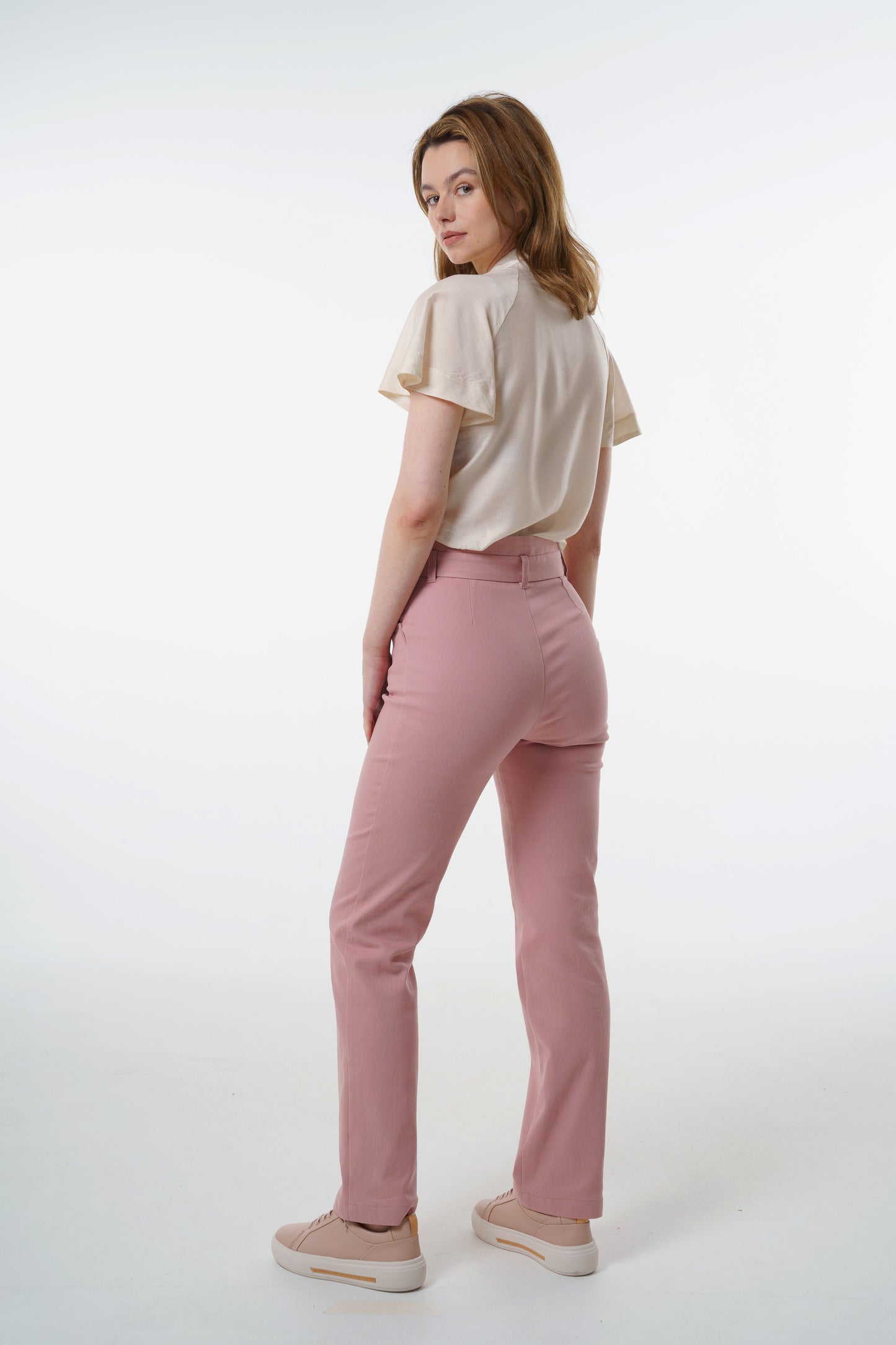 Rose‑dust high‑waisted stretch denim trousers with layered waistband and clean back seams, worn by a model, angled studio view.