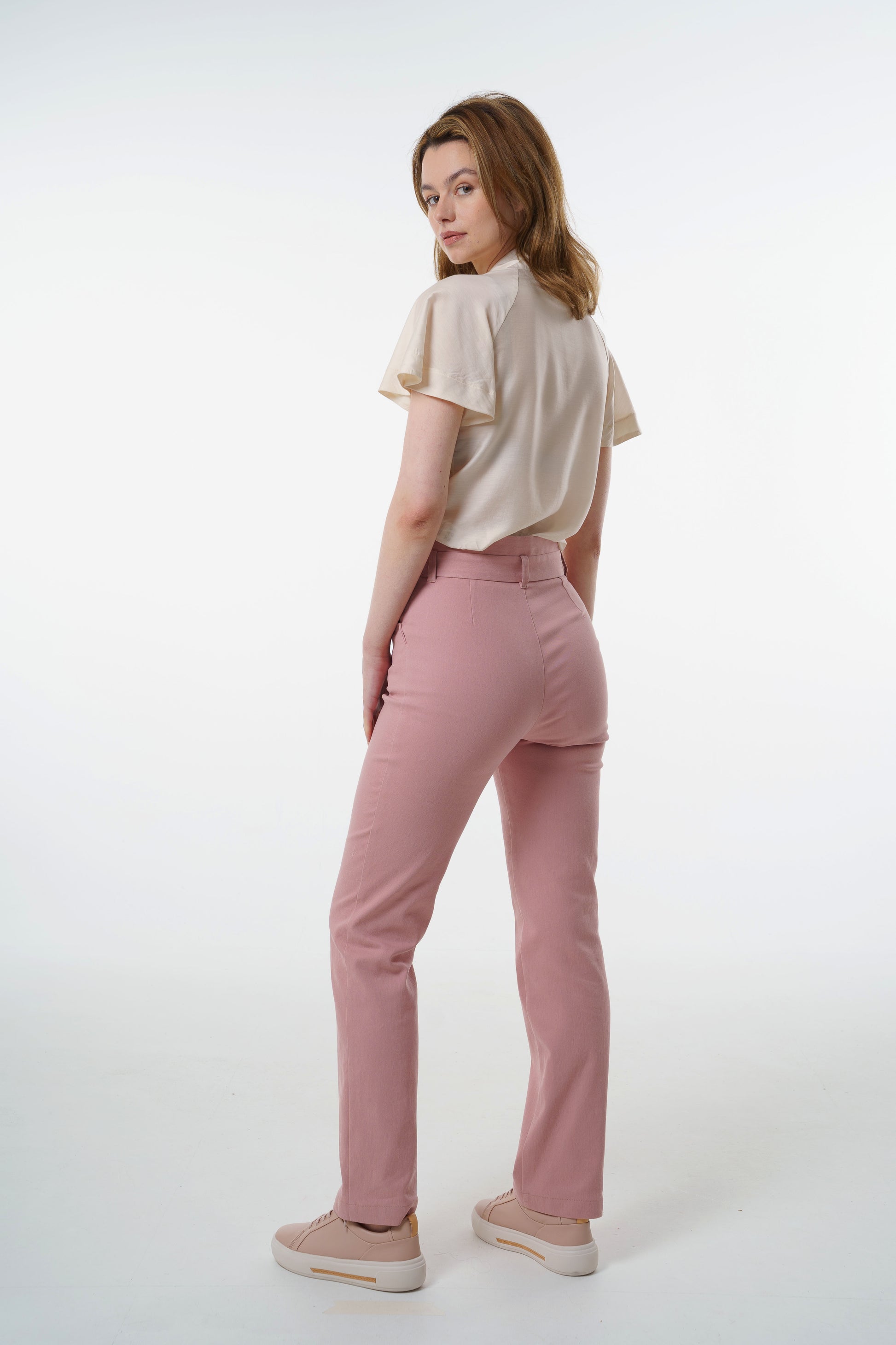 Rose‑dust high‑waisted stretch denim trousers with layered waistband and clean back seams, worn by a model, angled studio view.