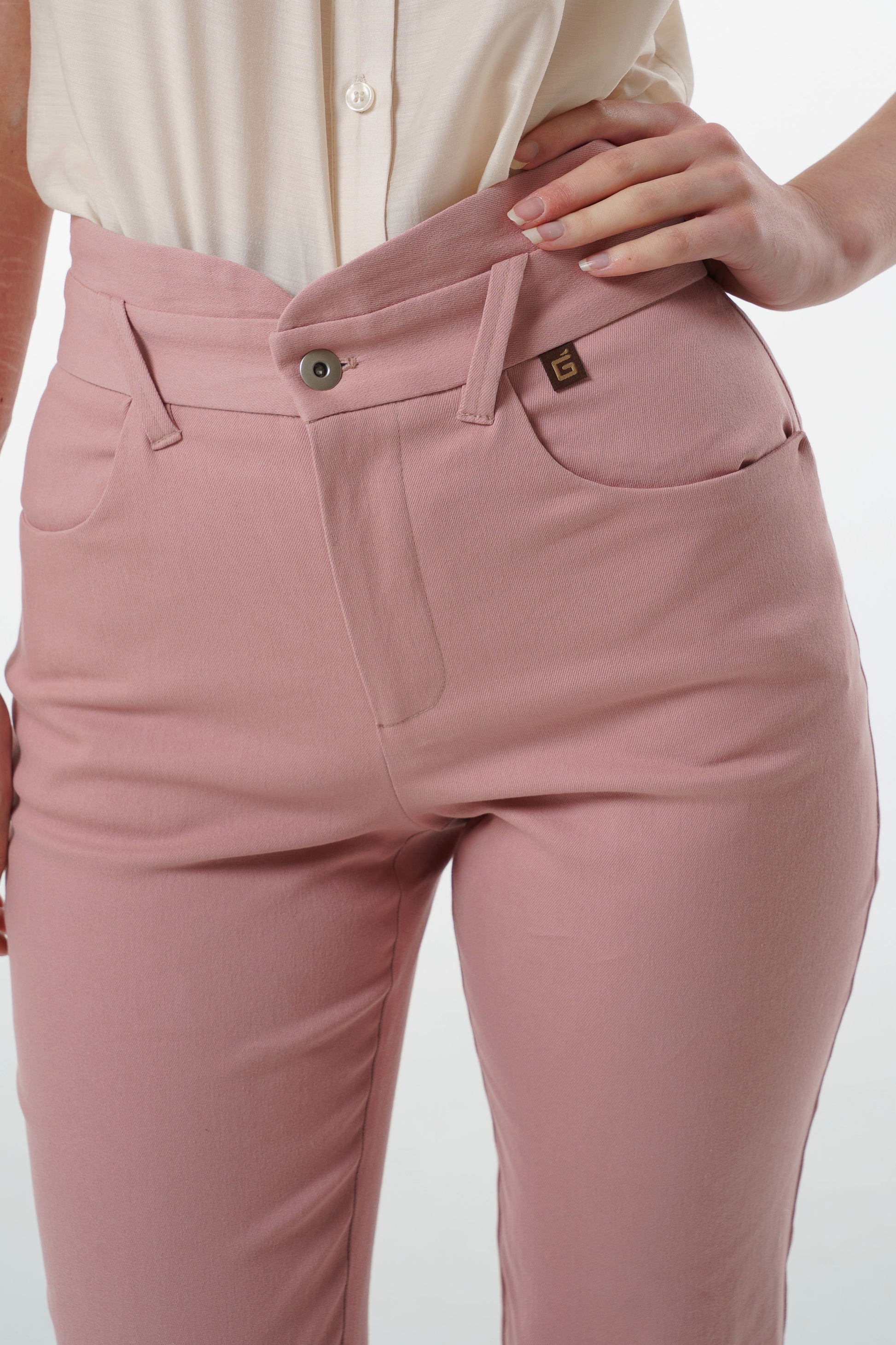 Close‑up of rose‑dust high‑waisted stretch denim trousers with layered waistband and front button closure, worn by a model.