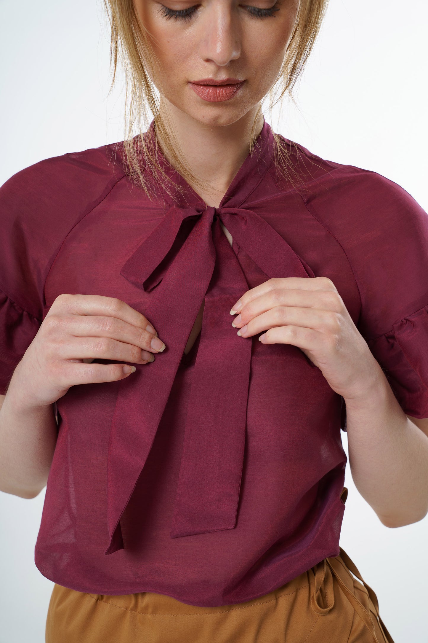 Close‑up of burgundy viscose–silk raglan‑sleeve blouse showing the neck tie and ruffled cuffs, worn by a model, studio detail view.