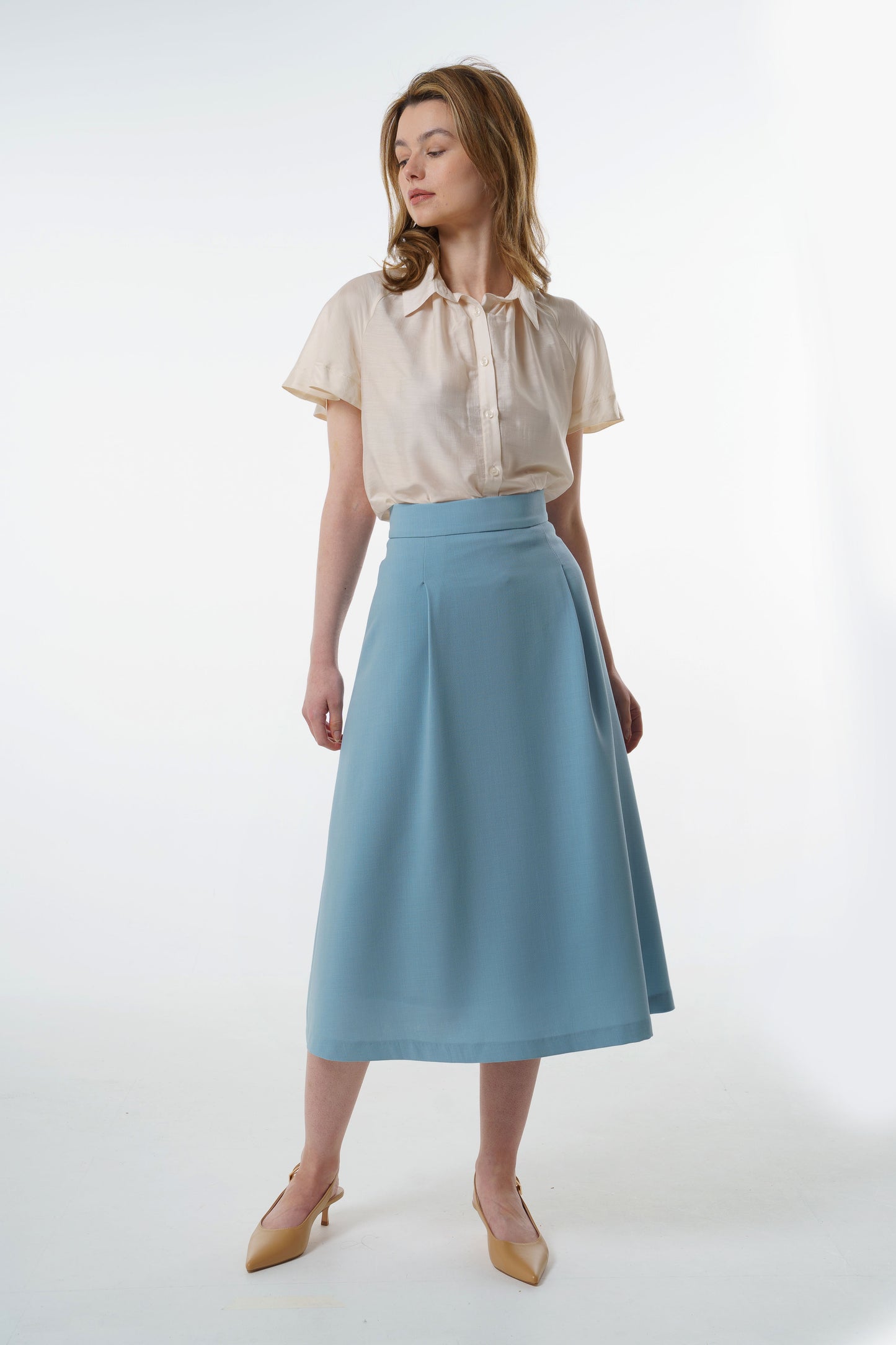 Light blue high‑waisted A‑line midi skirt in a virgin wool blend with clean flare and front pleat, worn by a model, full‑length studio view.