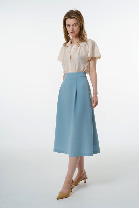 Light blue high‑waisted A‑line midi skirt in virgin wool with structured front pleat, worn by a model, front studio shot.