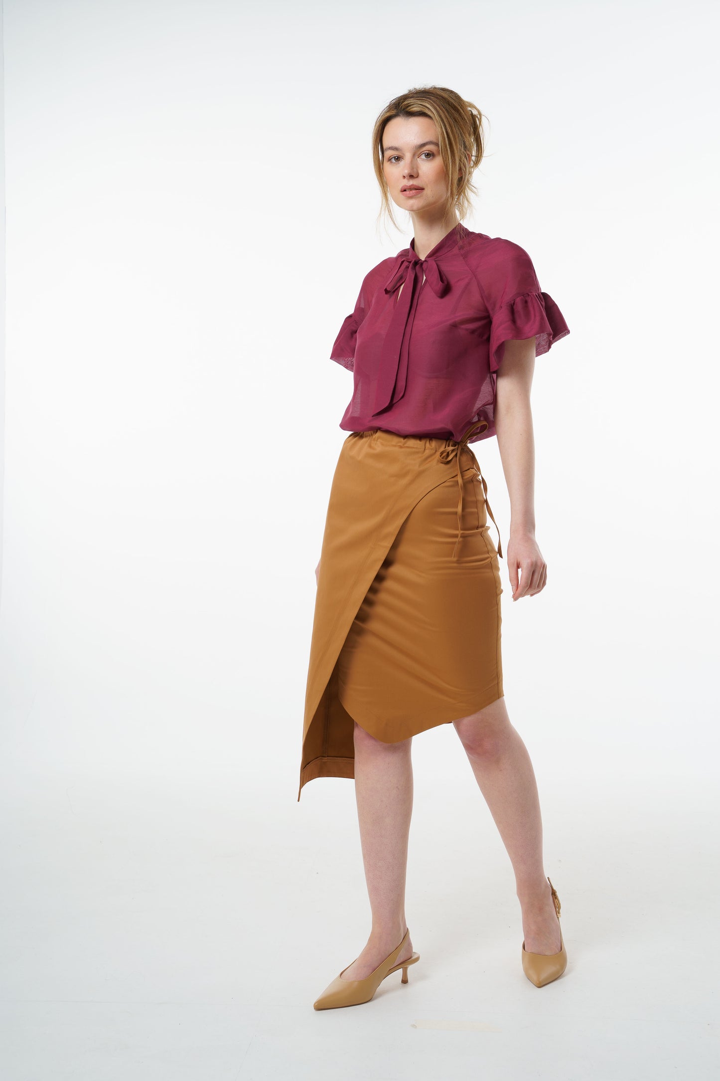 Camel cotton–viscose wrap pencil skirt with asymmetric overlay and high‑waisted fit, worn by a model, angled studio view.