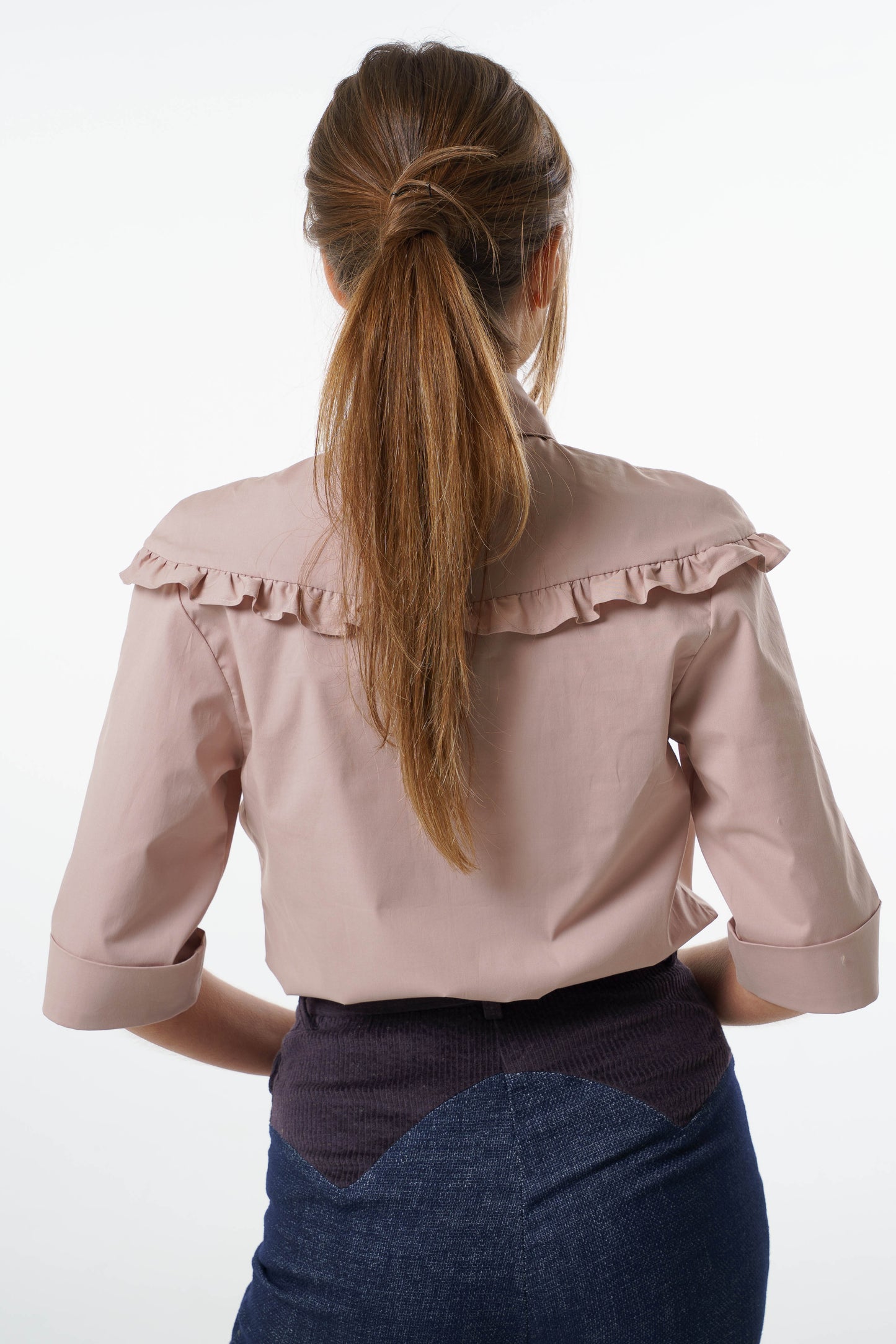 Blush cotton‑stretch shirt with ruffled back yoke and three‑quarter sleeves, worn by a model with high‑waisted two‑tone trousers, back studio view.