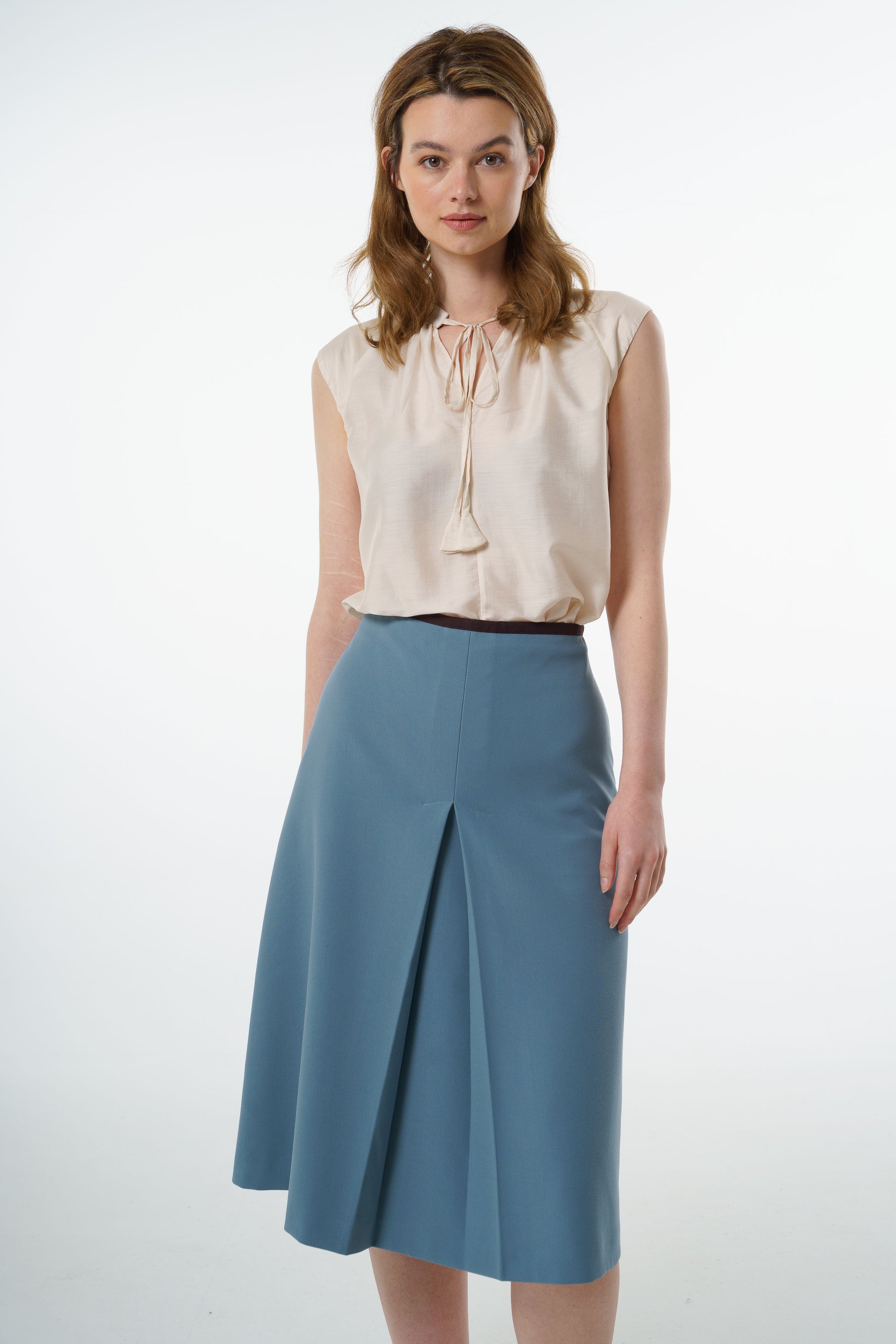 Ivory sleeveless silk–viscose blouse with front tie and fluid drape, worn by a model with a blue A‑line skirt, front studio shot.
