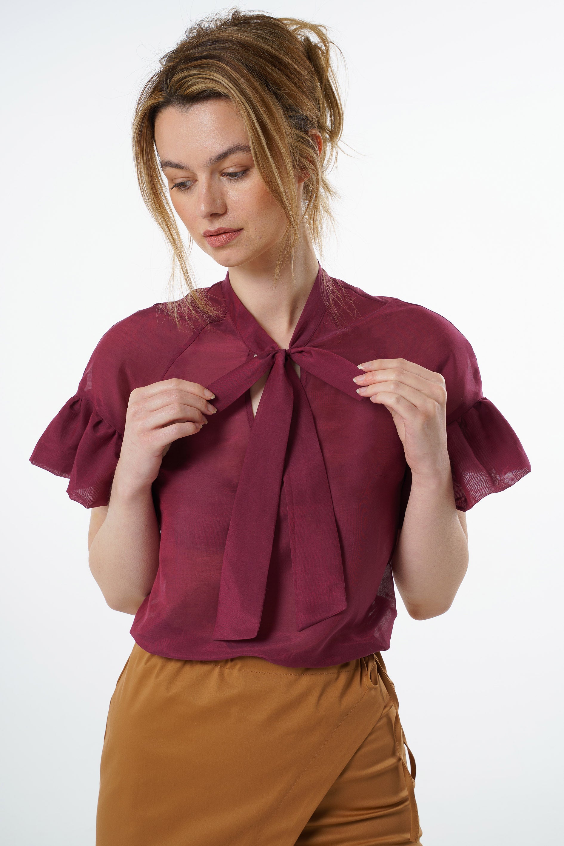 Burgundy viscose–silk raglan‑sleeve blouse with neck tie and ruffled cuffs, worn by a model with high‑waisted mustard trousers, close‑up studio view.