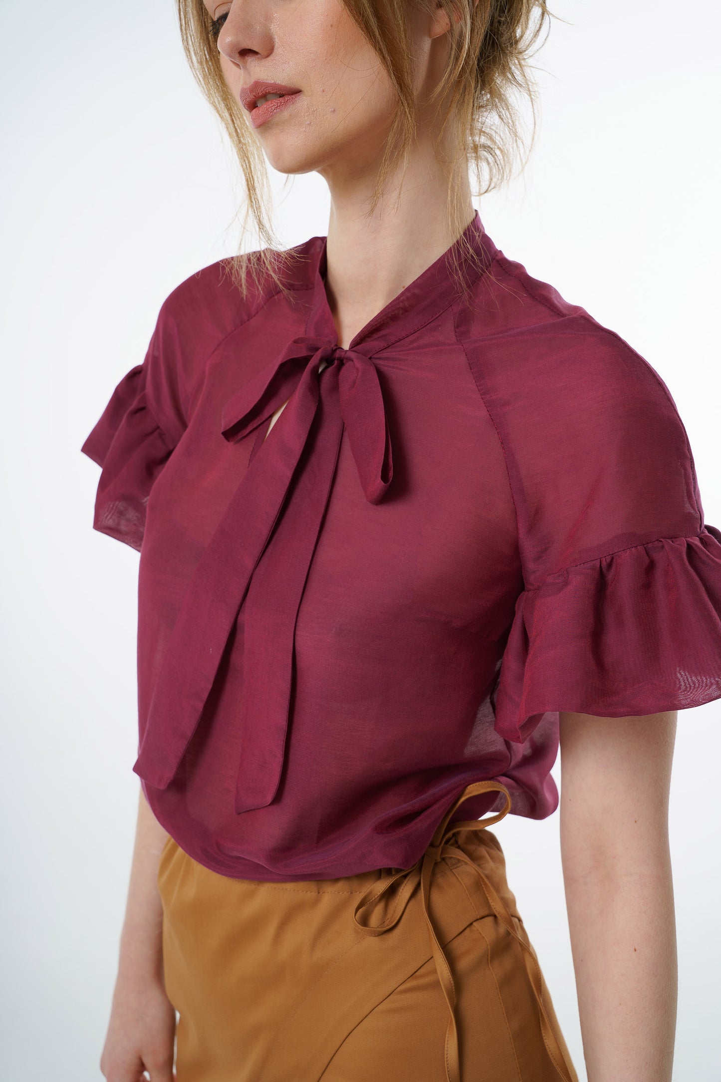 Burgundy viscose–silk raglan‑sleeve blouse with neck tie and ruffled cuffs, worn by a model with a mustard wrap skirt, angled studio view.