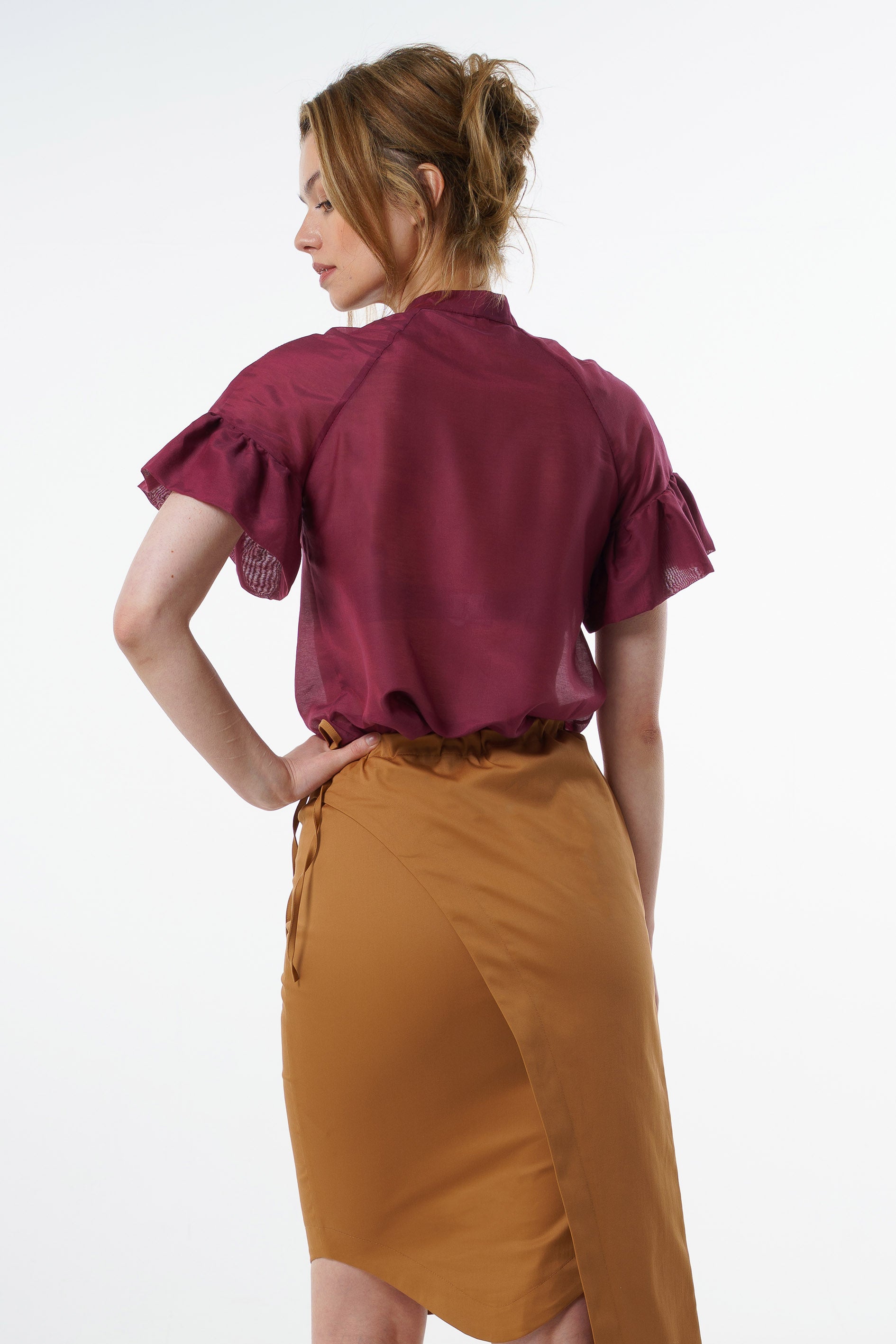 Burgundy viscose–silk raglan‑sleeve blouse with ruffled cuffs and soft back drape, worn by a model with a mustard wrap skirt, back studio view.