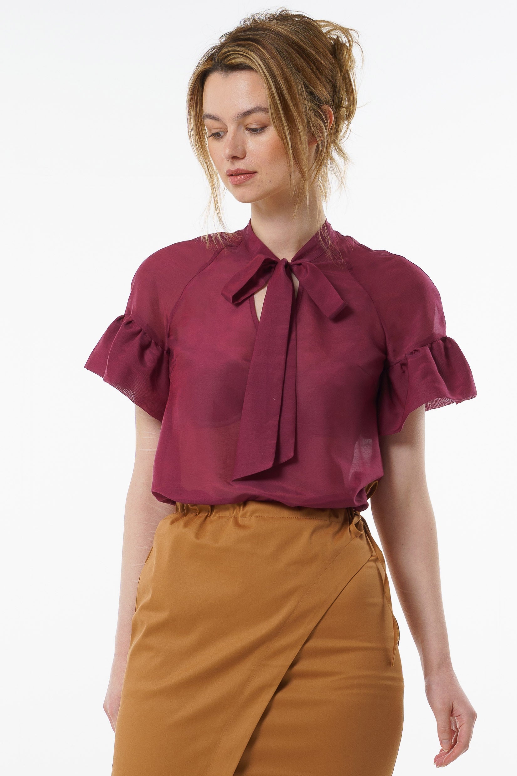Burgundy viscose–silk raglan‑sleeve blouse with neck tie and ruffled cuffs, worn by a model with high‑waisted mustard trousers, front studio view.