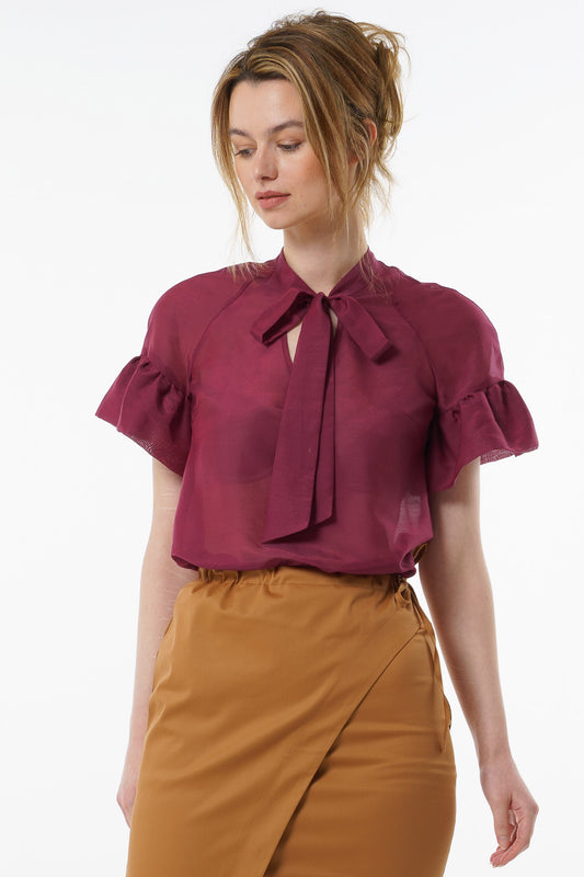 Burgundy viscose–silk raglan‑sleeve blouse with neck tie and ruffled cuffs, worn by a model with high‑waisted mustard trousers, front studio view.