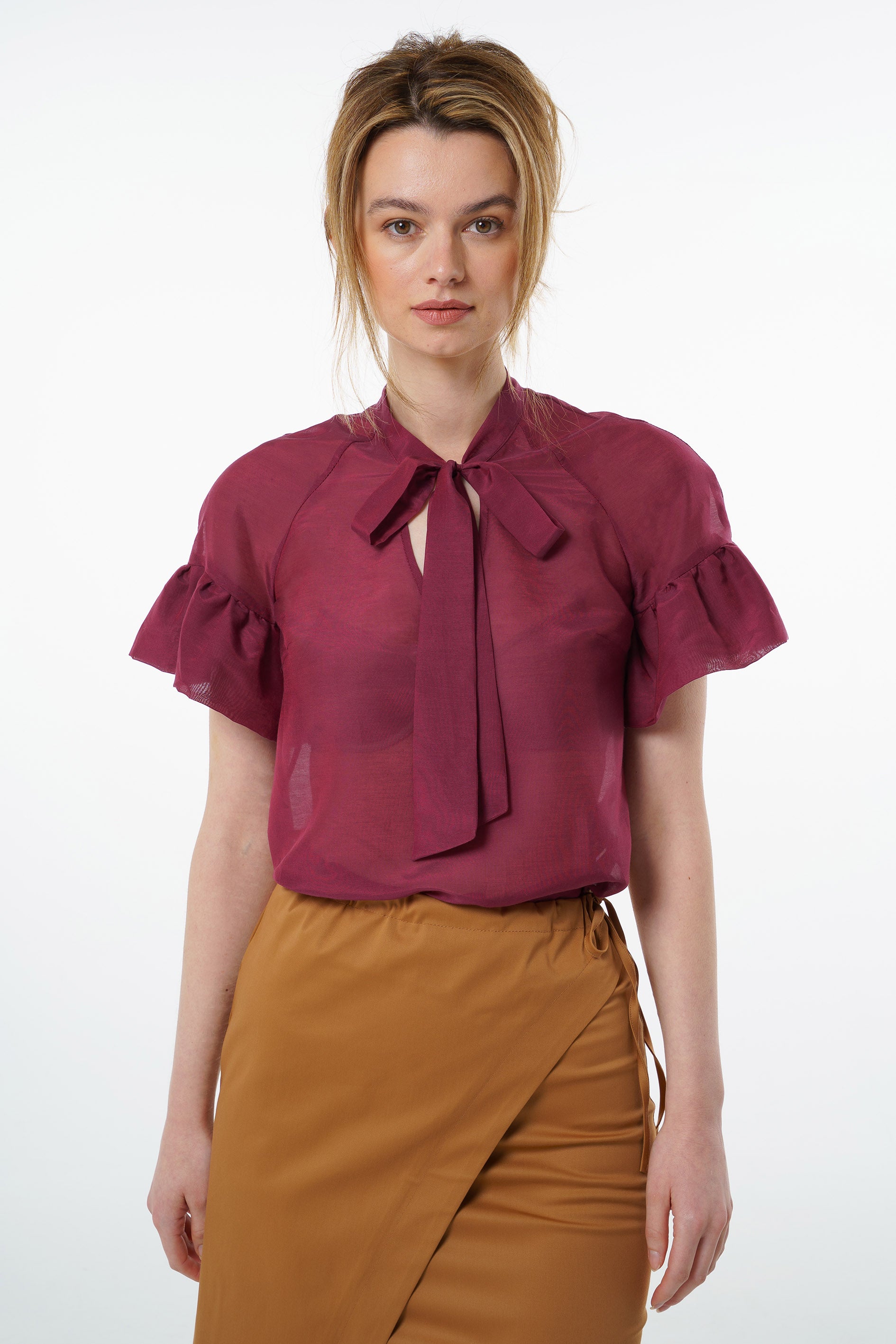 Burgundy viscose–silk raglan‑sleeve blouse with neck tie and soft ruffled cuffs, worn by a model with a high‑waisted mustard wrap skirt, front studio view.