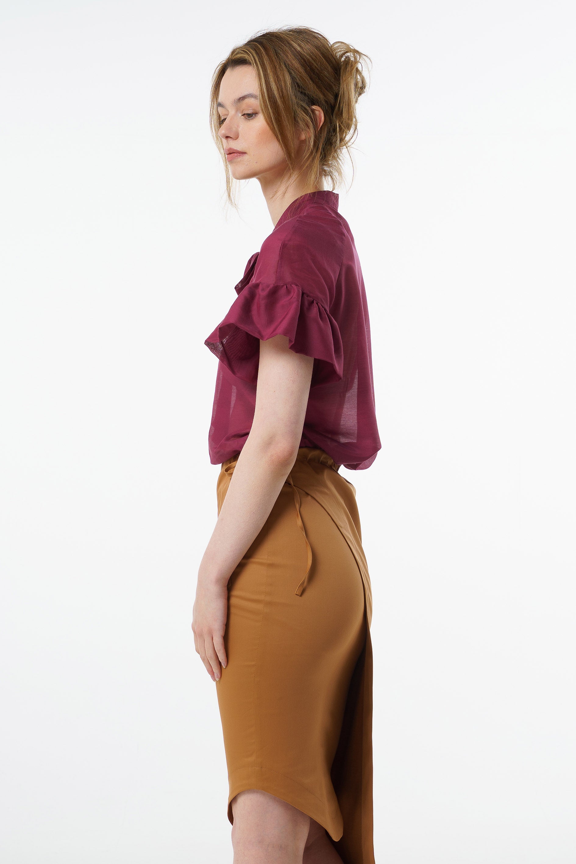 Burgundy viscose–silk raglan‑sleeve blouse with neck tie and ruffled cuffs, worn by a model with a high‑waisted mustard skirt, side studio view.