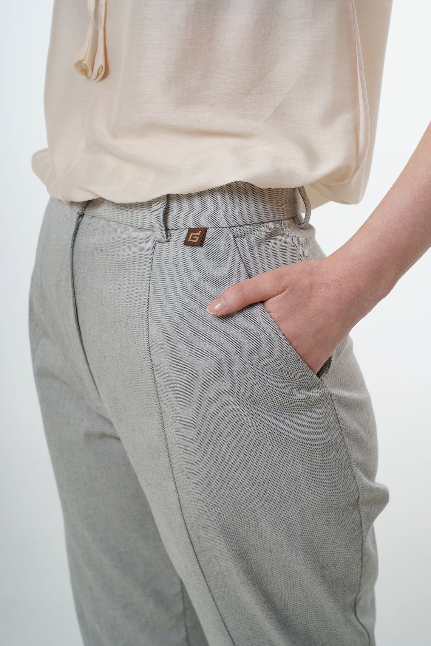 Close‑up of light grey tapered carrot‑leg trousers in a cotton blend showing high‑waisted construction, front pocket, and subtle logo tab, worn by a model.