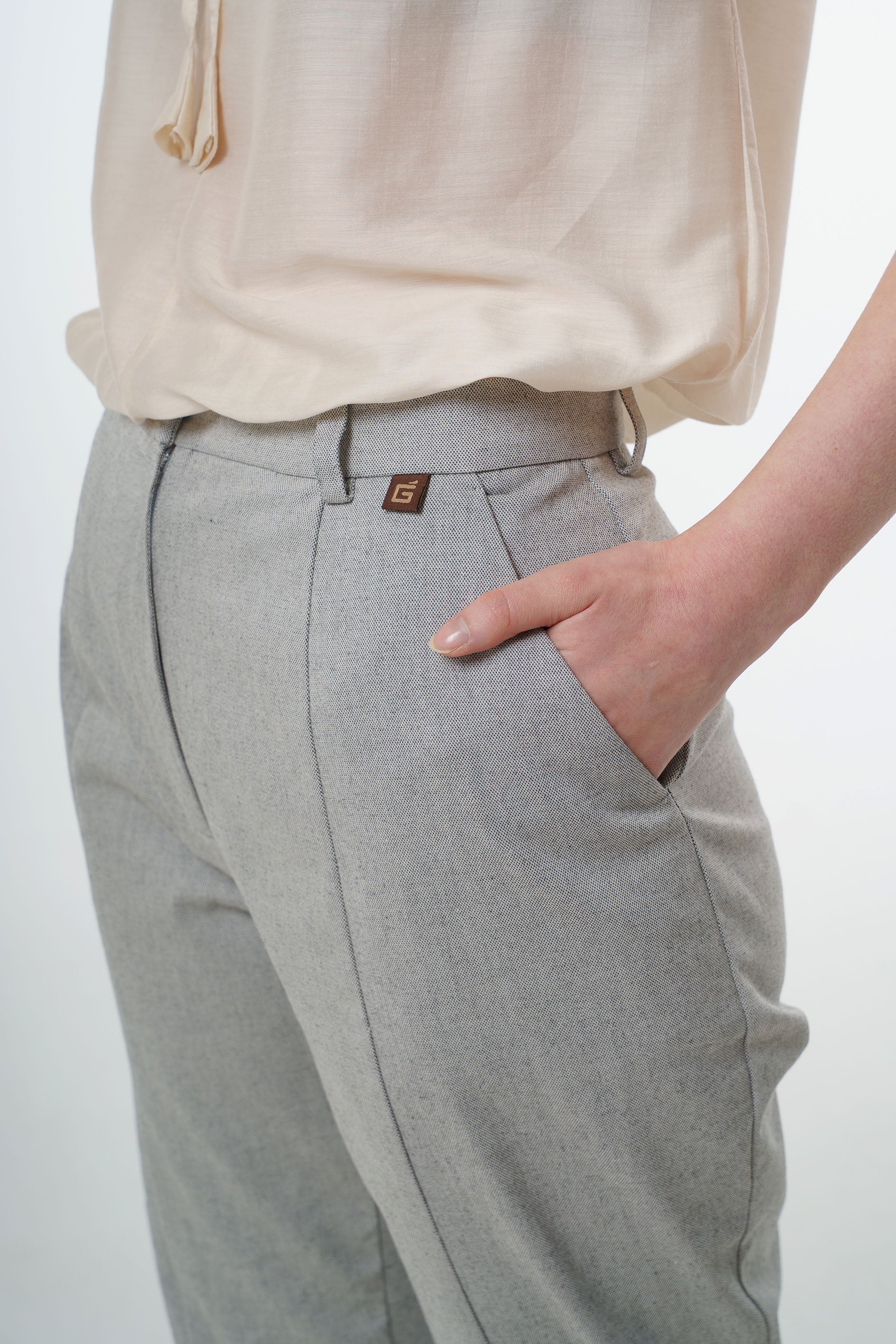 Close‑up of light grey tapered carrot‑leg trousers in a cotton blend showing high‑waisted construction, front pocket, and subtle logo tab, worn by a model.