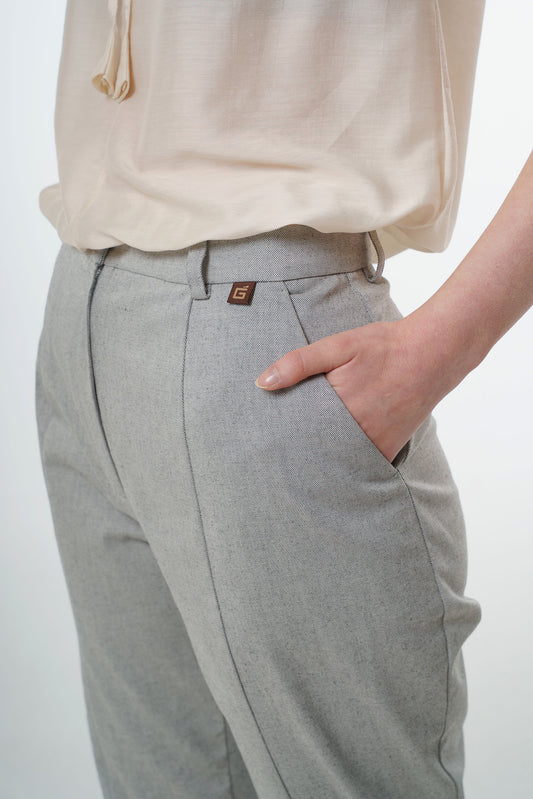 Close‑up of light grey tapered carrot‑leg trousers in a cotton blend showing high‑waisted construction, front pocket, and subtle logo tab, worn by a model.