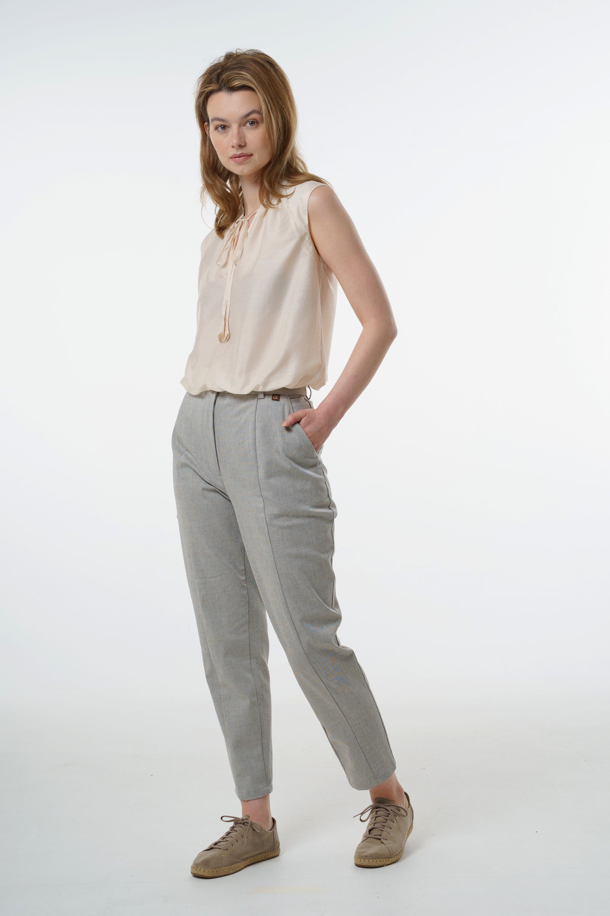 Light grey tapered carrot‑leg trousers in a cotton blend with front seam detailing and high‑waisted fit, worn by a model with a sleeveless blouse, front studio view.