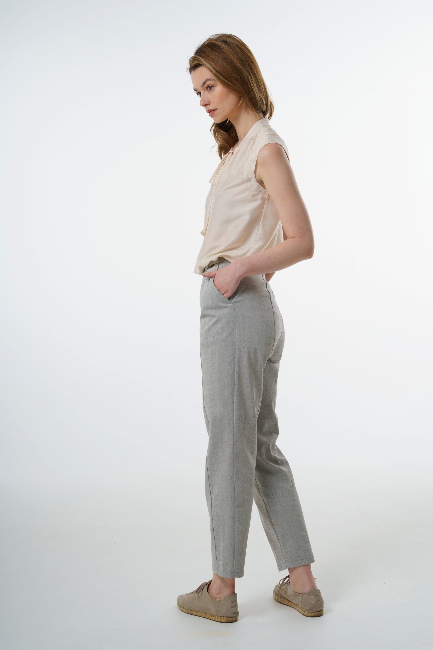 - Light grey tapered carrot‑leg trousers in a cotton blend with clean side line and high‑waisted fit, worn by a model, side studio view.