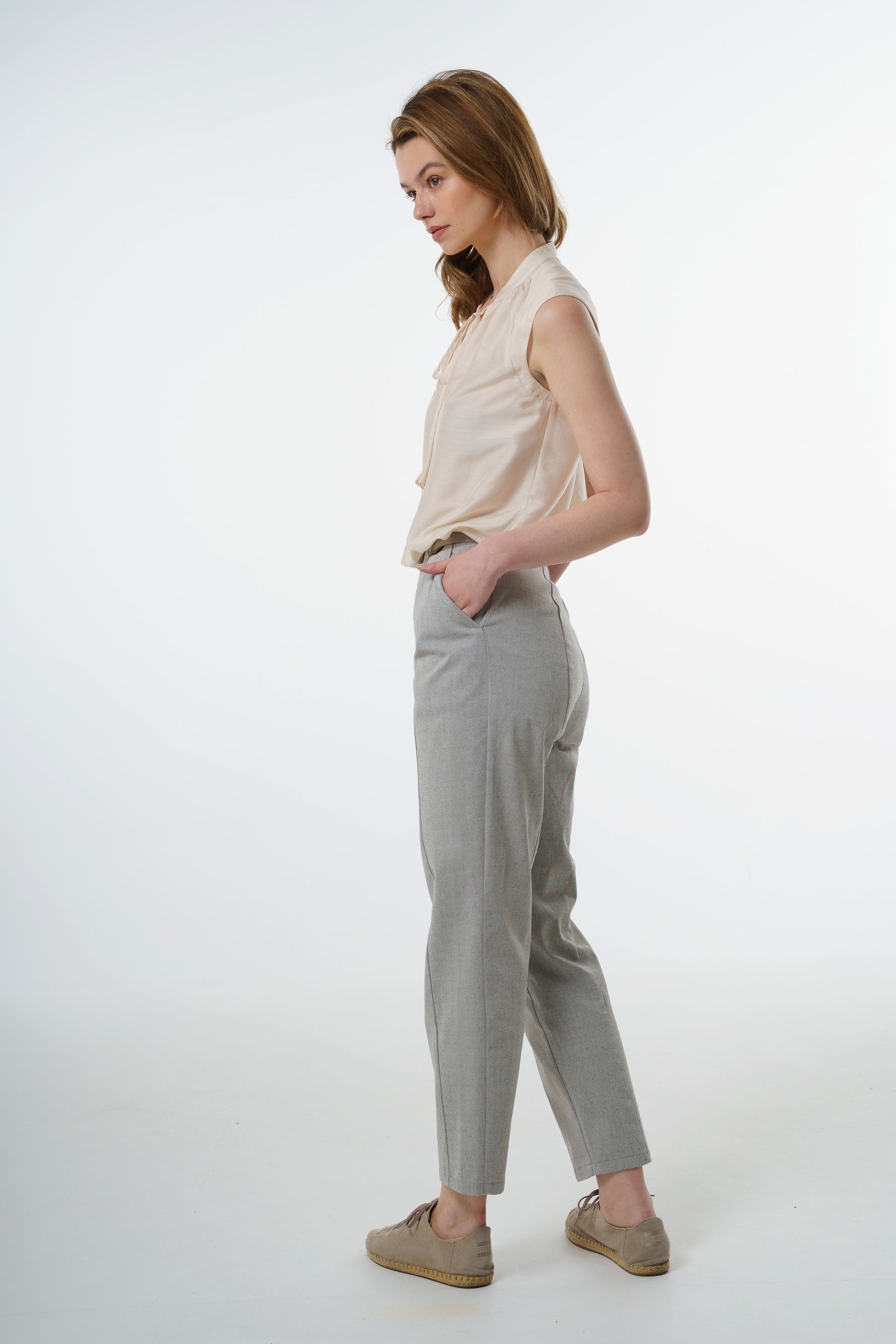 - Light grey tapered carrot‑leg trousers in a cotton blend with clean side line and high‑waisted fit, worn by a model, side studio view.